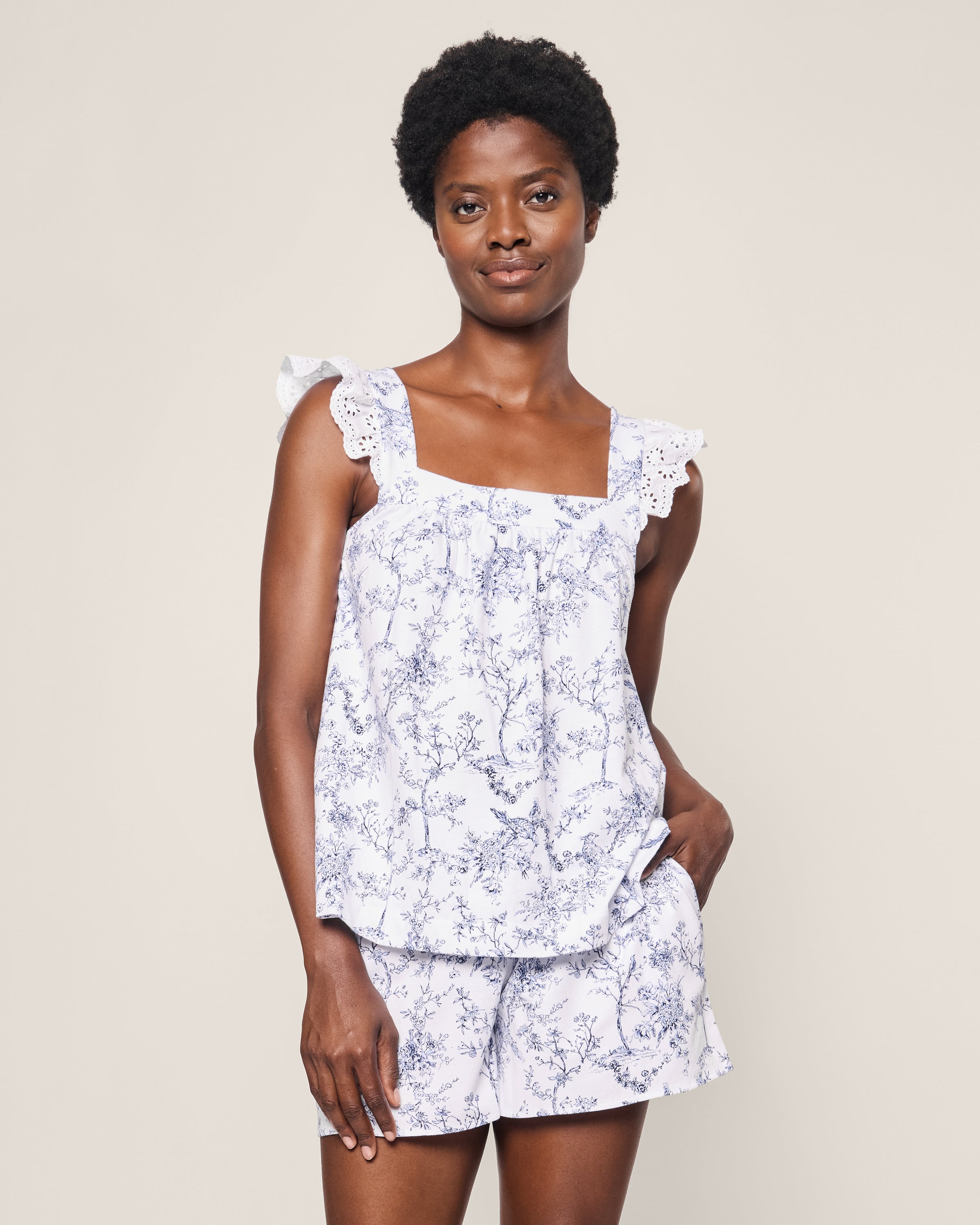 Wearing the Womens Pima Clara Short Set in Timeless Toile by Petite Plume, featuring a blue floral pattern and ruffled shoulder straps, one exudes botanical elegance with their short curly hair against a plain background.