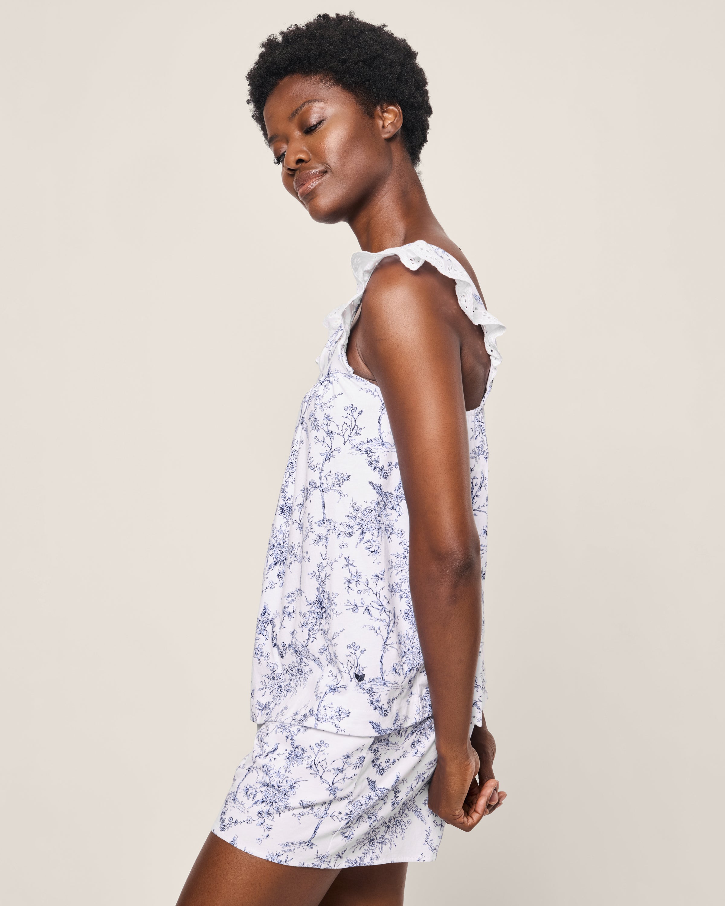 Wearing Petite Plumes Womens Pima Clara Short Set in Timeless Toile, a person stands in profile against a plain background. The ruffled shoulders and blue floral patterns on the white fabric exude botanical elegance as they look down with a gentle smile.