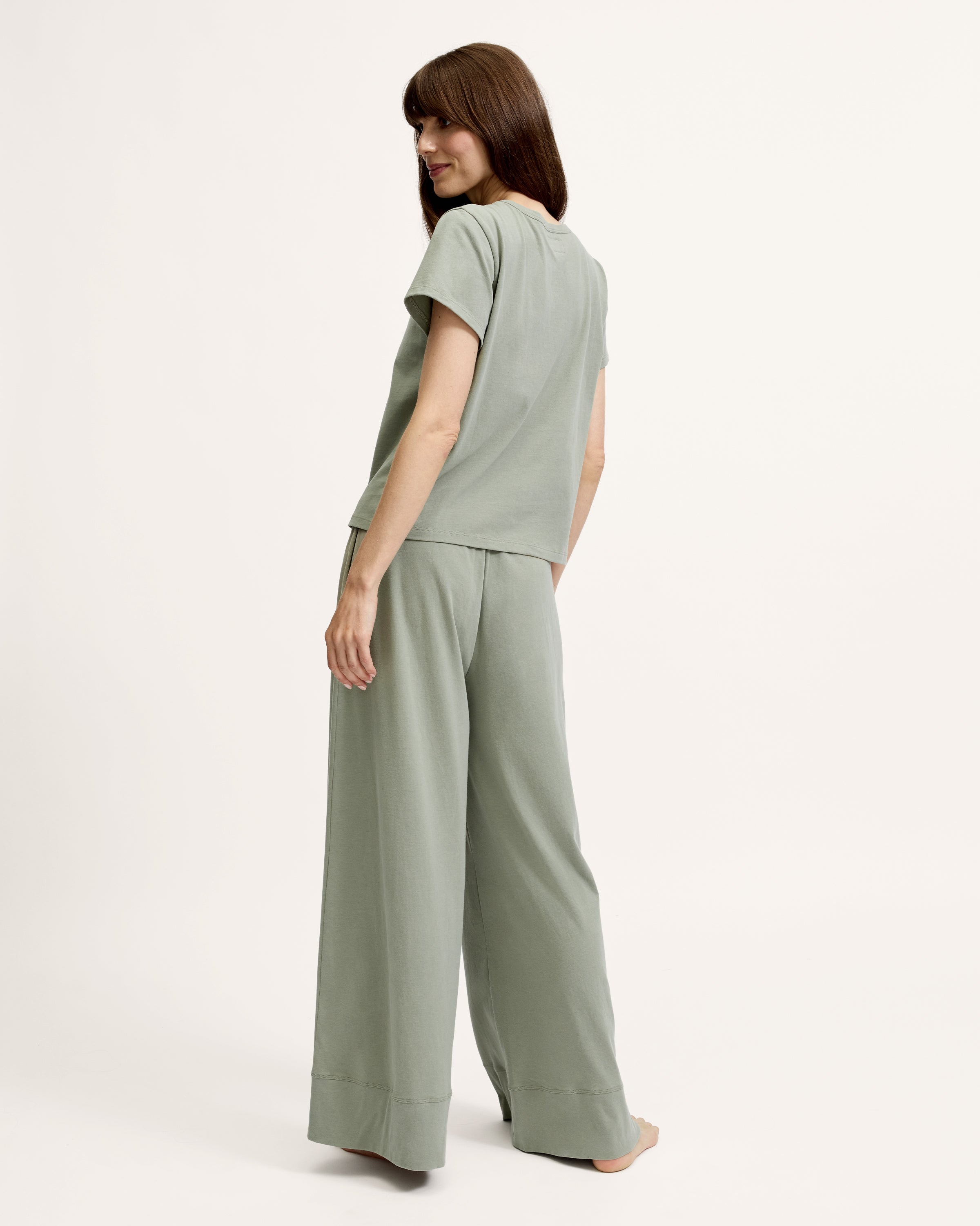 A woman with straight brown hair wears the Women's Pima Essential Crew Neck Tee in Sage and matching drawstring shorts, standing with her hands on her hips against a plain light background.
