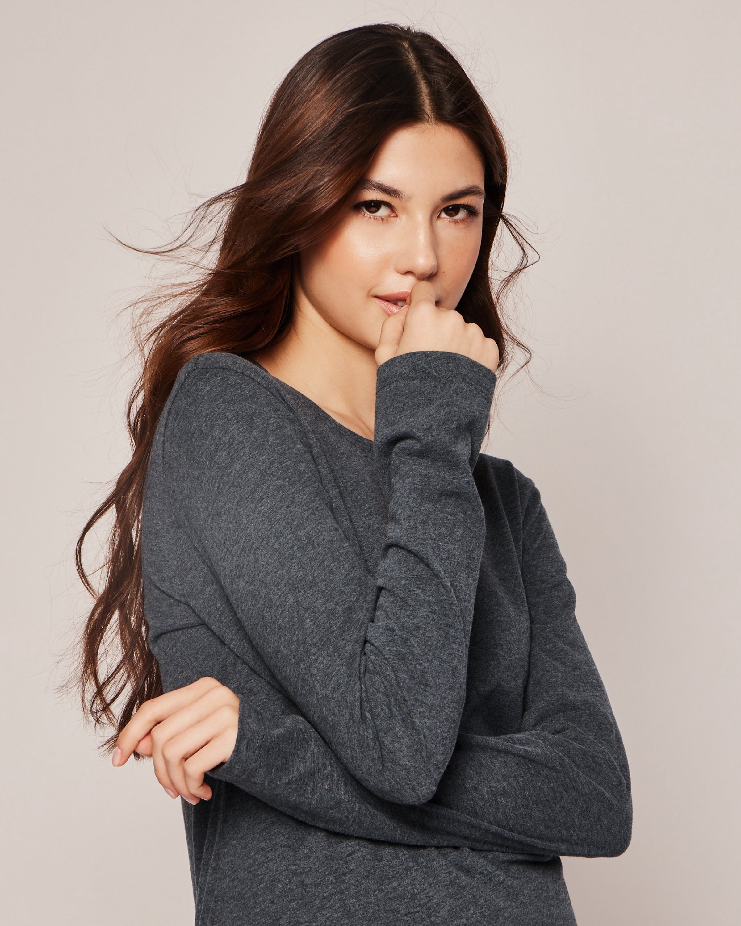 A person with long, dark hair wears Petite Plumes Womens Pima Long Sleeve Top in Dark Heather Grey. They look thoughtfully at the camera, one hand near their mouth and the other on their arm against a light backdrop. Crafted from luxurious Peruvian Pima cotton.