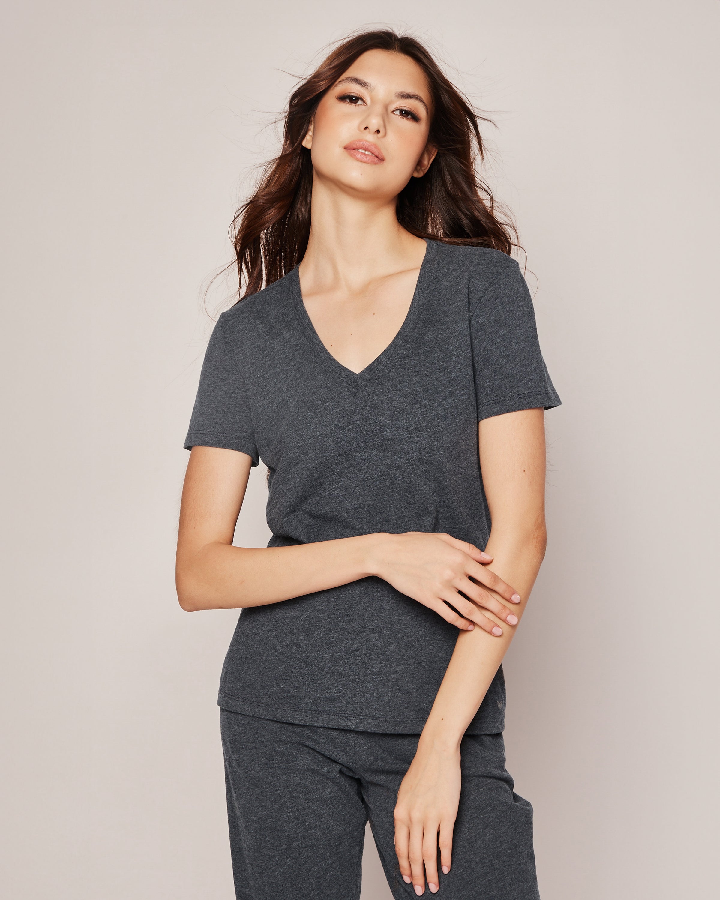 A person with long hair poses in luxurious Petite Plume pajamas made from breathable fabric. The Womens Pima Short Sleeve Top in Dark Heather Grey complements the neutral background, with one hand on their waist and the other crossed over.