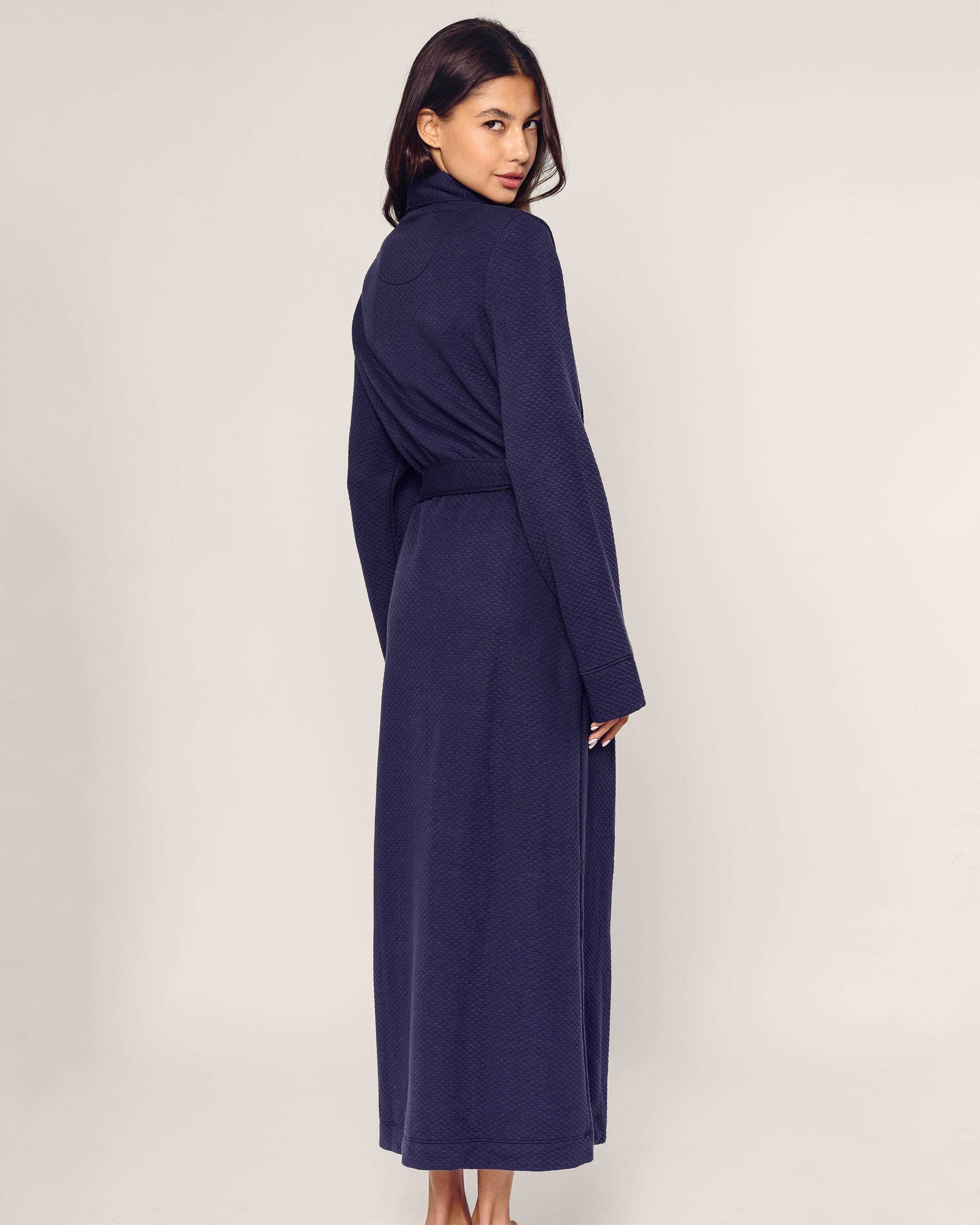 A person stands with their back turned, glancing over their shoulder, wearing the Womens Pima Ophelia Robe in Blue Dusk Jacquard by Petite Plume. The plain, light-colored background highlights the robes elegant quilted effect.