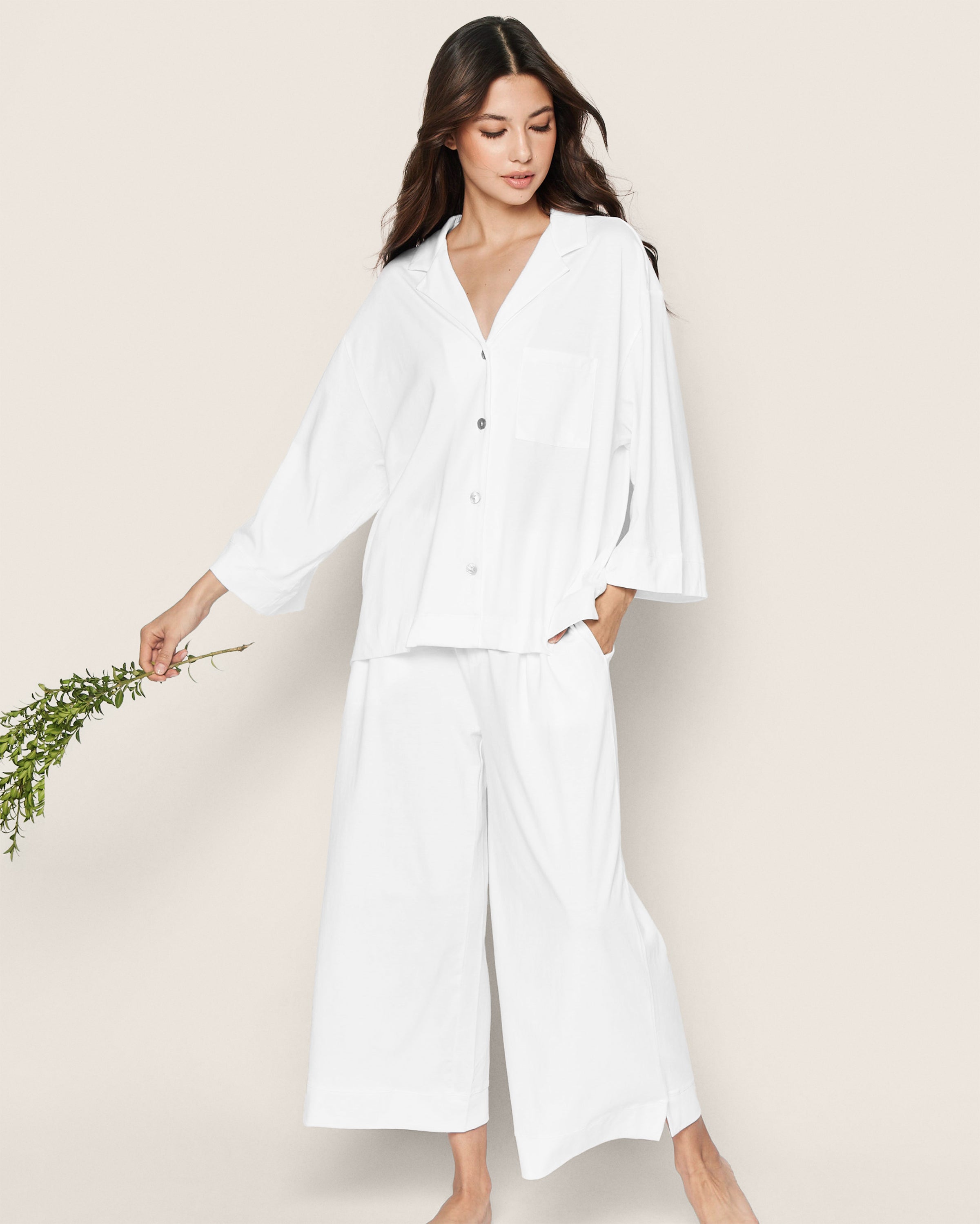 Barefoot and holding a branch with leaves, a person dons the loose-fitting Olivia Wide Leg Pima Pajama Set in White with Mama Embroidery by Petite Plume. The button-up shirt and pants against a light background capture the essence of a special edition moment ideal for Mothers Day.