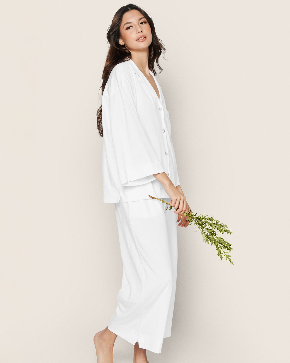 Wearing the Greek Embroidered Olivia Wide Leg Pima Pajama Set in White, a woman with long dark hair stands barefoot against a beige background, gently holding a small bouquet of green foliage and displaying a relaxed, serene expression.