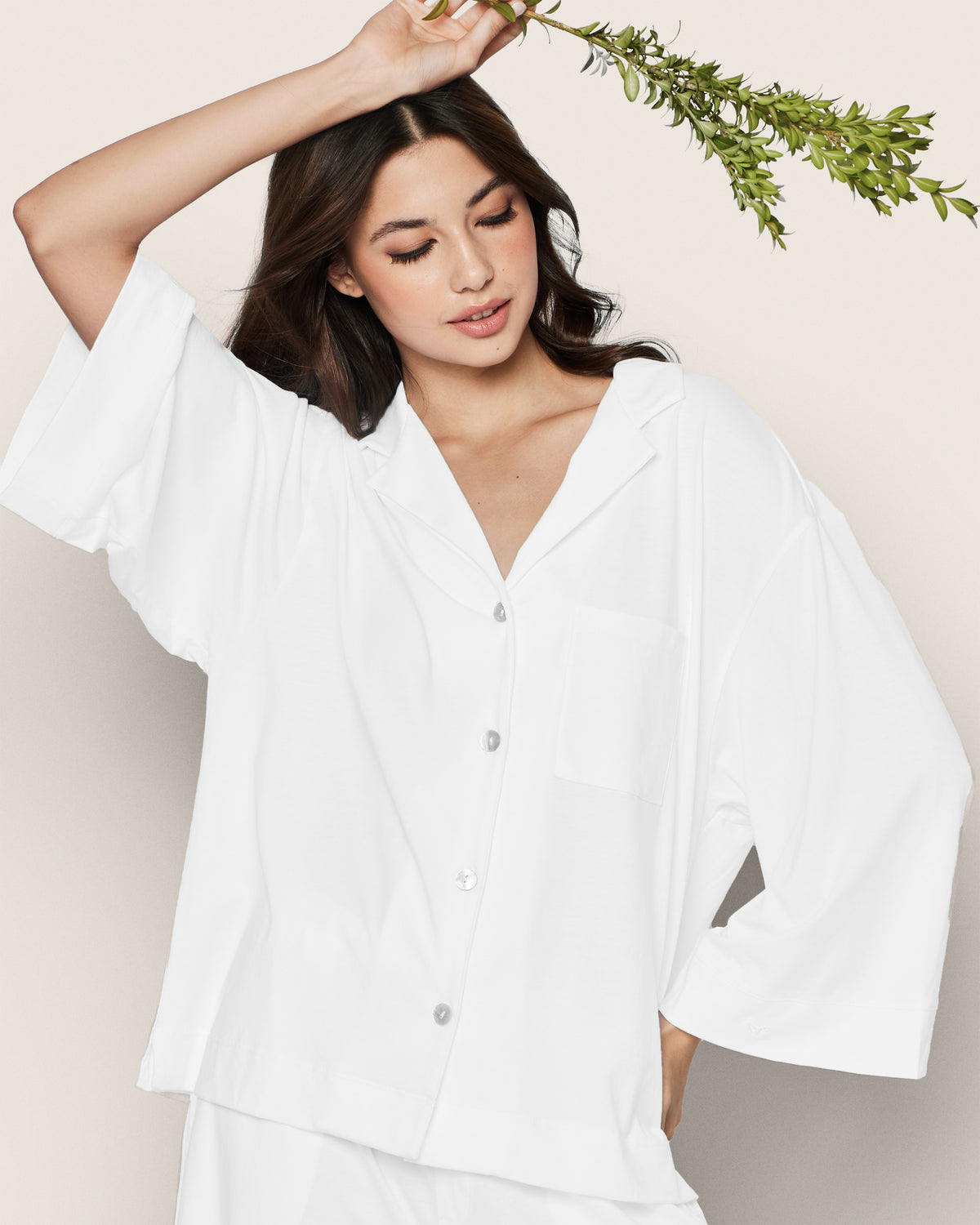 A woman wearing the Greek Embroidered Olivia Wide Leg Pima Pajama Set in White poses with one arm raised, holding a green leafy branch, in front of a neutral background.