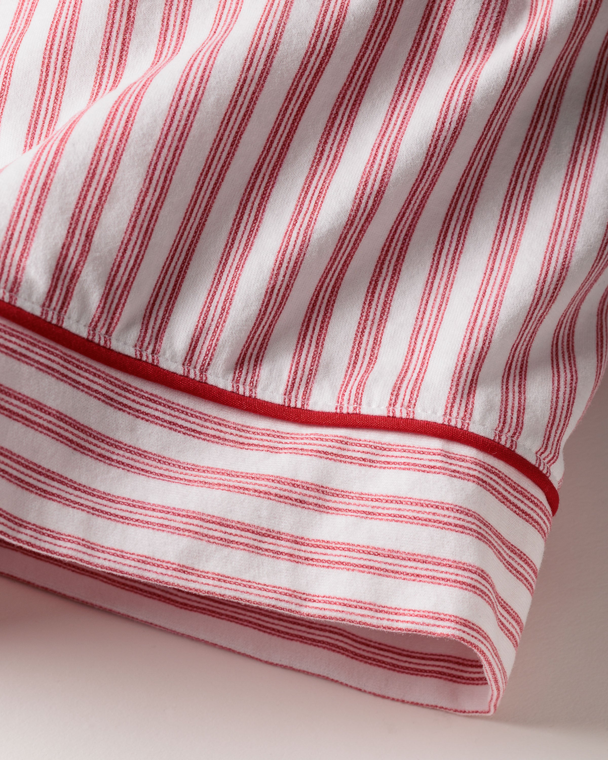 A close-up of Petite Plumes Womens Pima Long Sleeve Short Set in Antique Red Ticking reveals a Pima cotton fabric with consistent red and white vertical stripes, tailored piping, and a solid red band at the bottom. The fabric rests on a light surface, casting a soft shadow.