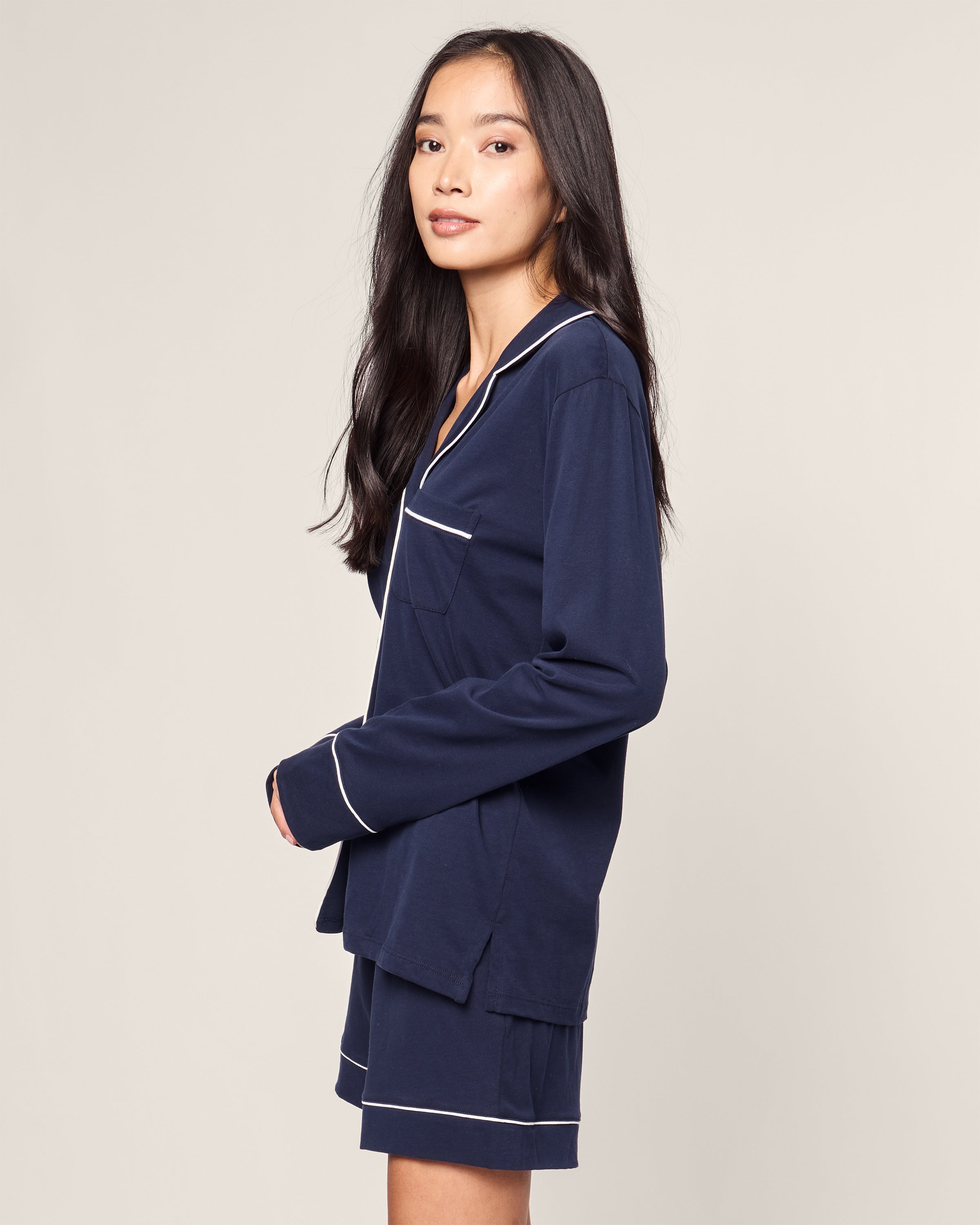 A person with long, dark hair is wearing the Petite Plume Womens Pima Long Sleeve Short Set in Navy, featuring white piping, and standing sideways against a plain background.