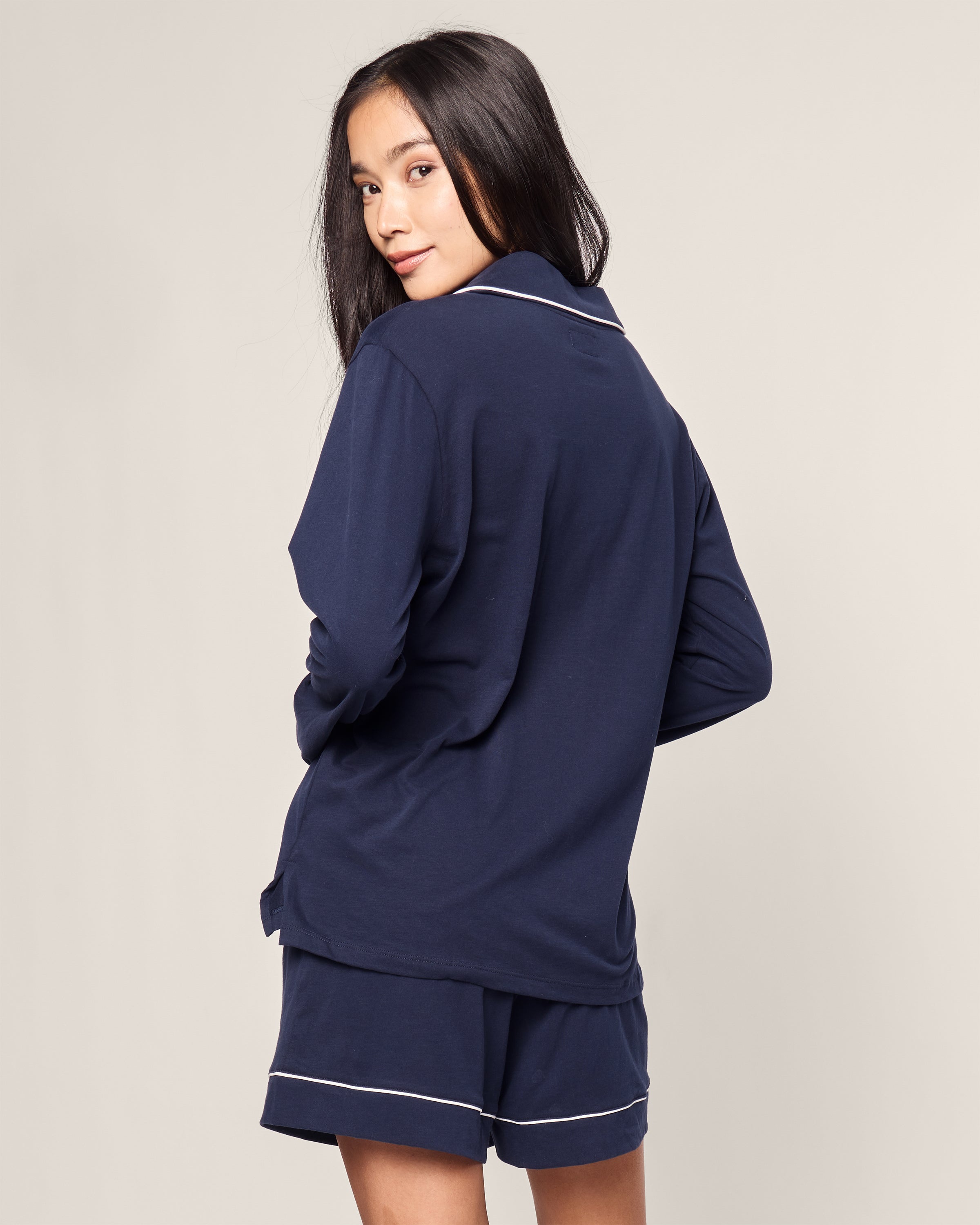 A woman with long dark hair, wearing the Womens Pima Long Sleeve Short Set in Navy by Petite Plume, stands sideways and looks back over her shoulder against a neutral background.