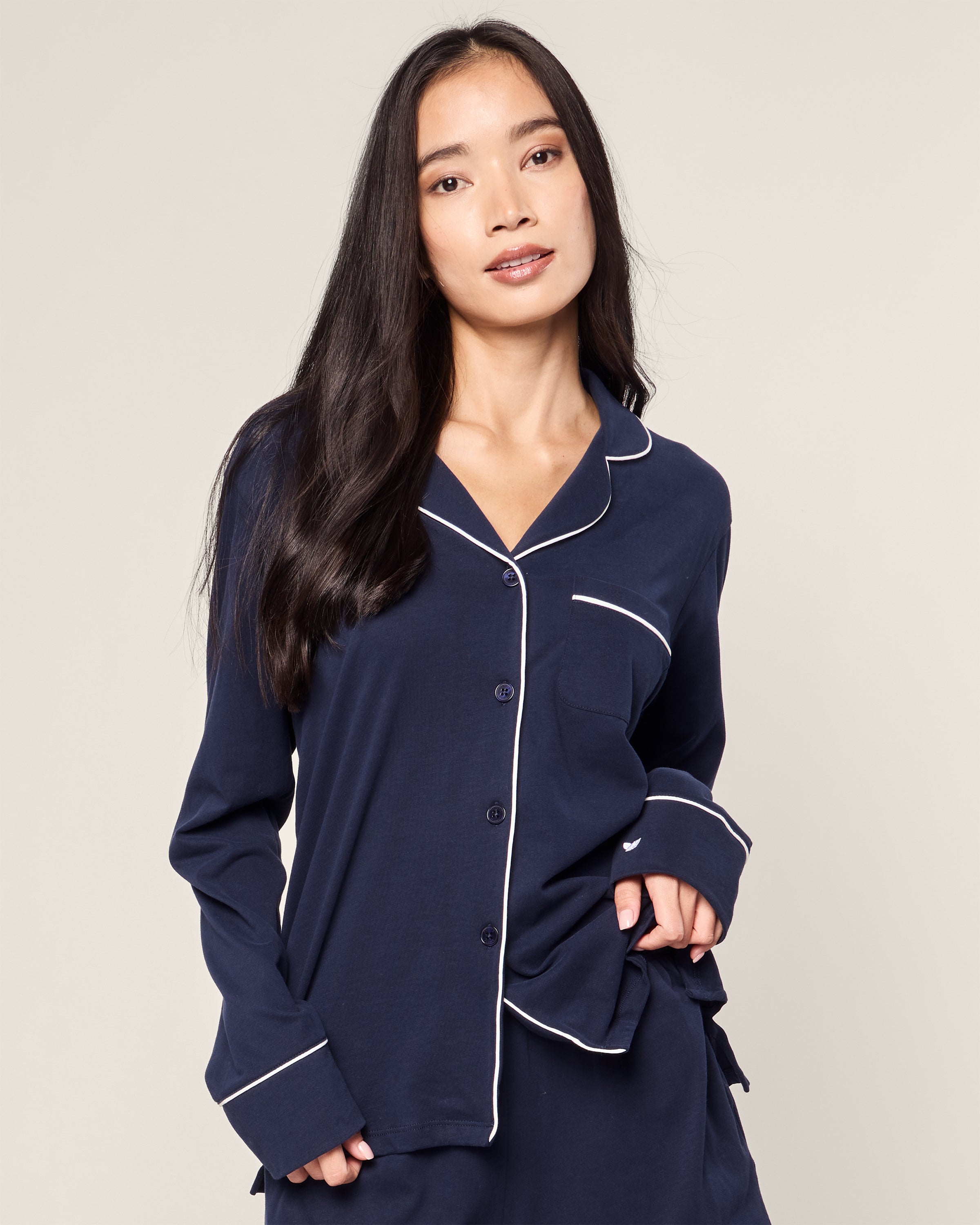 A woman with long dark hair wears Petite Plumes Womens Pima Long Sleeve Short Set in Navy, crafted from luxurious Peruvian Pima cotton with white piping. Her relaxed posture and gentle expression embody the essence of Classic PJs against a plain background.