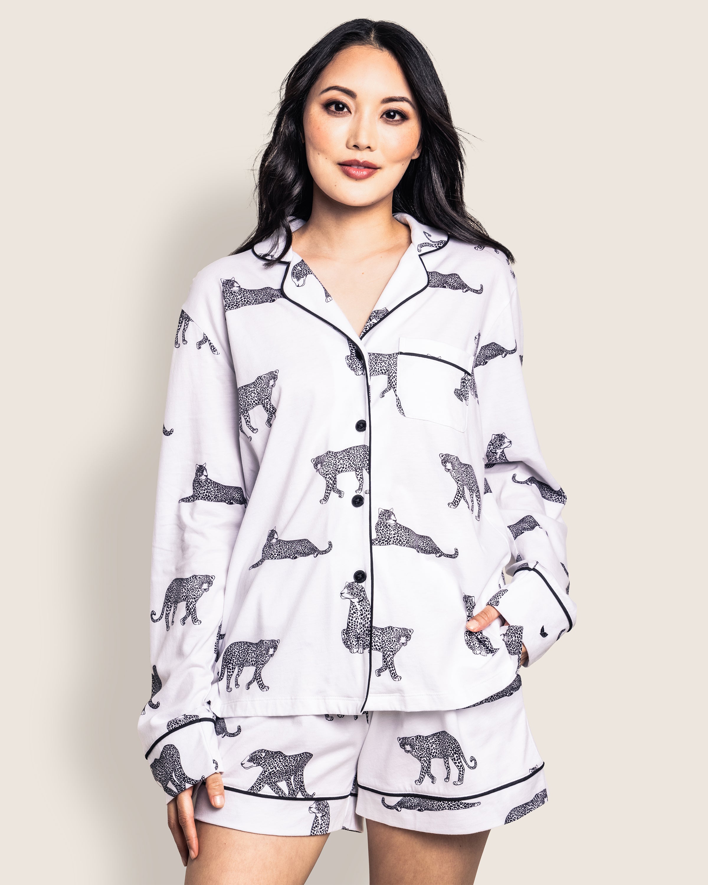 A woman models Petite Plumes Womens Pima Short Set in Panthère de Paris, confidently wearing the white luxury pajamas with black leopard print. Crafted from soft Peruvian Pima cotton, the set includes a long-sleeve shirt and shorts. Her hands rest casually in her pockets as she gazes at the camera.