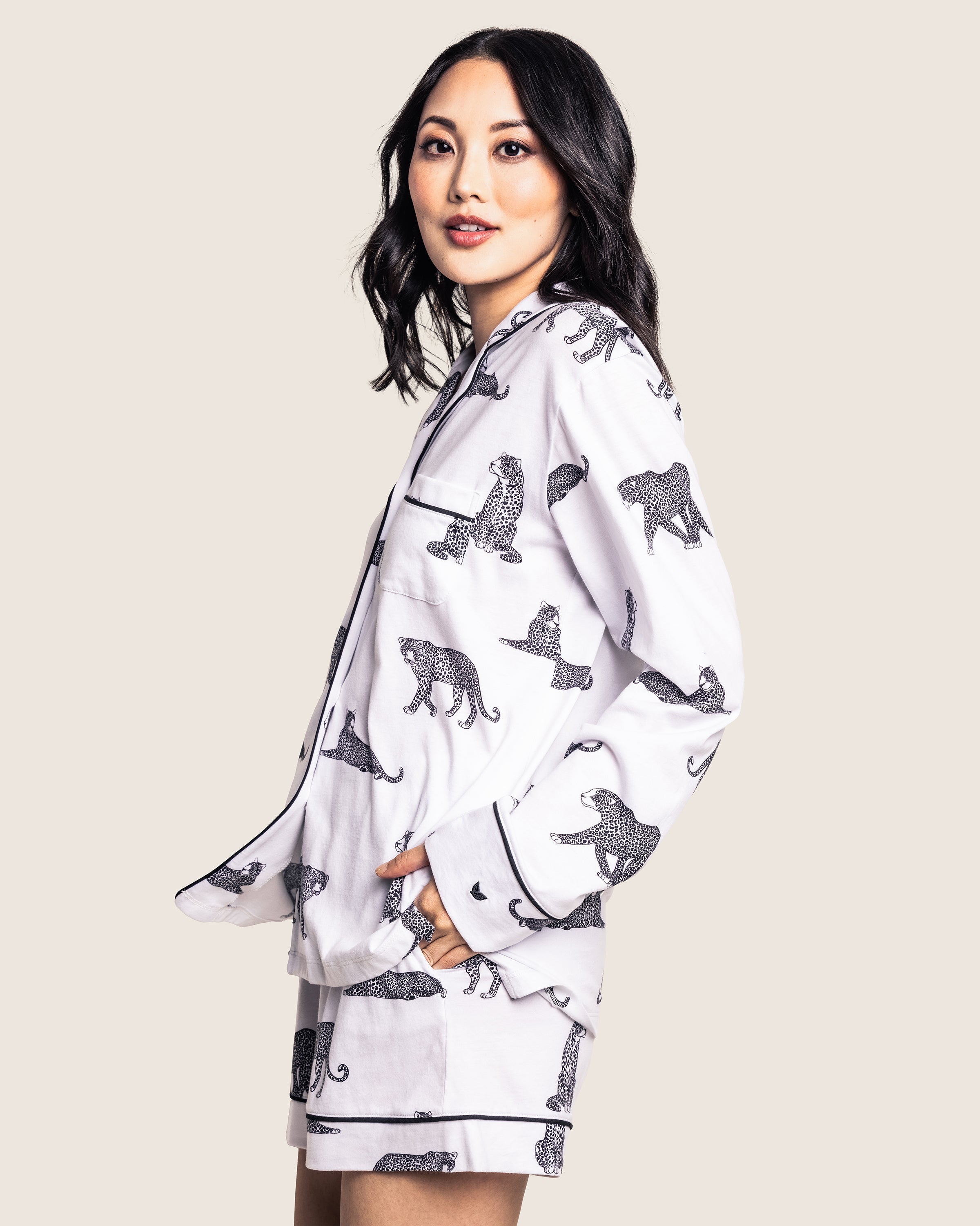 A person with long dark hair stands sideways in Petite Plumes Womens Pima Short Set in Panthère de Paris. This white pajama set, adorned with black leopard prints, is made from exquisite Luxe Pima cotton and includes a long-sleeve top and shorts against a plain light background, enhancing its luxury.