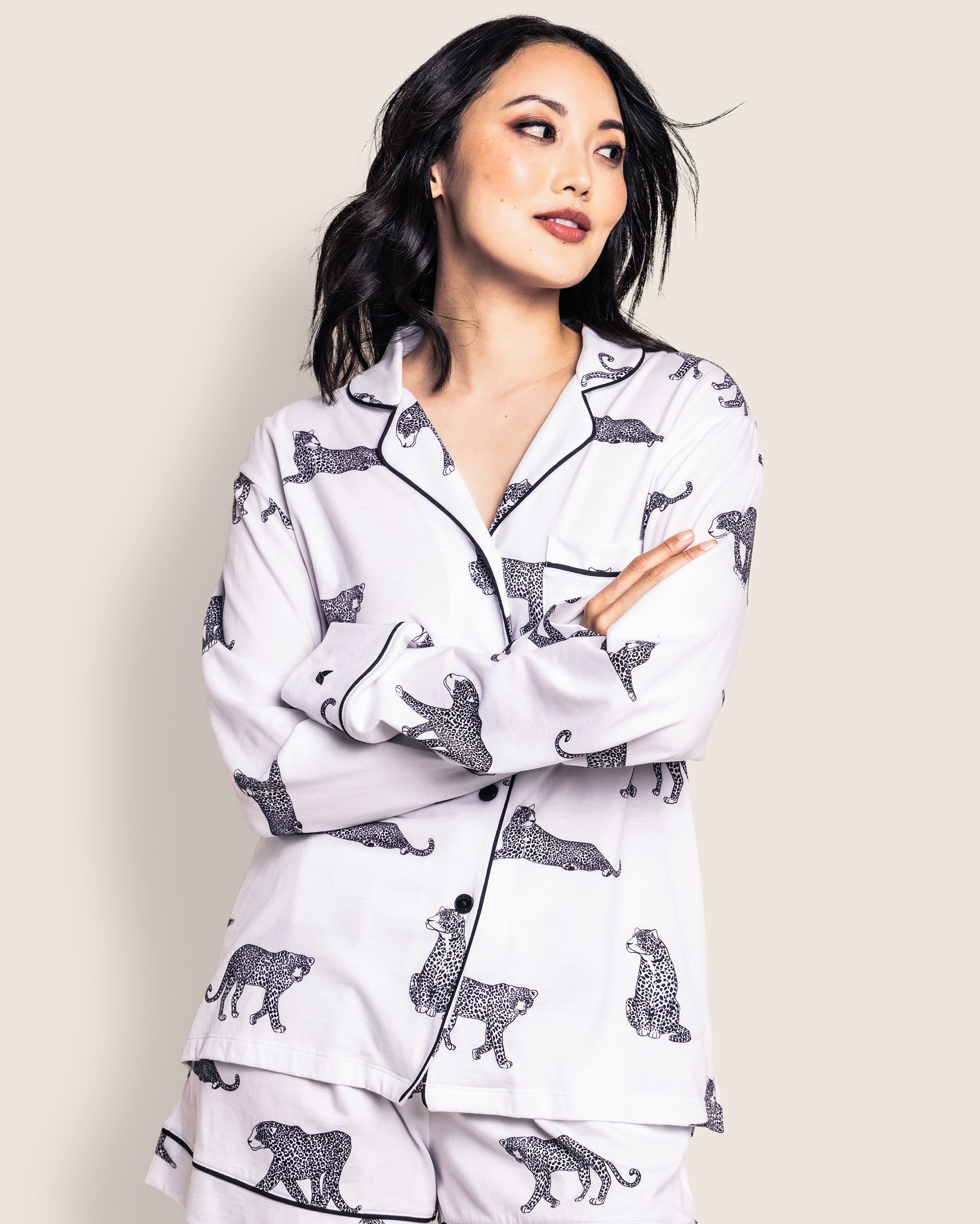 A person with long dark hair stands with folded arms, exuding effortless elegance in the Womens Pima Short Set in Panthère de Paris by Petite Plume. Their neutral gaze is to the side, against a light beige background, while luxury Peruvian Pima cotton displays a chic black cheetah print.