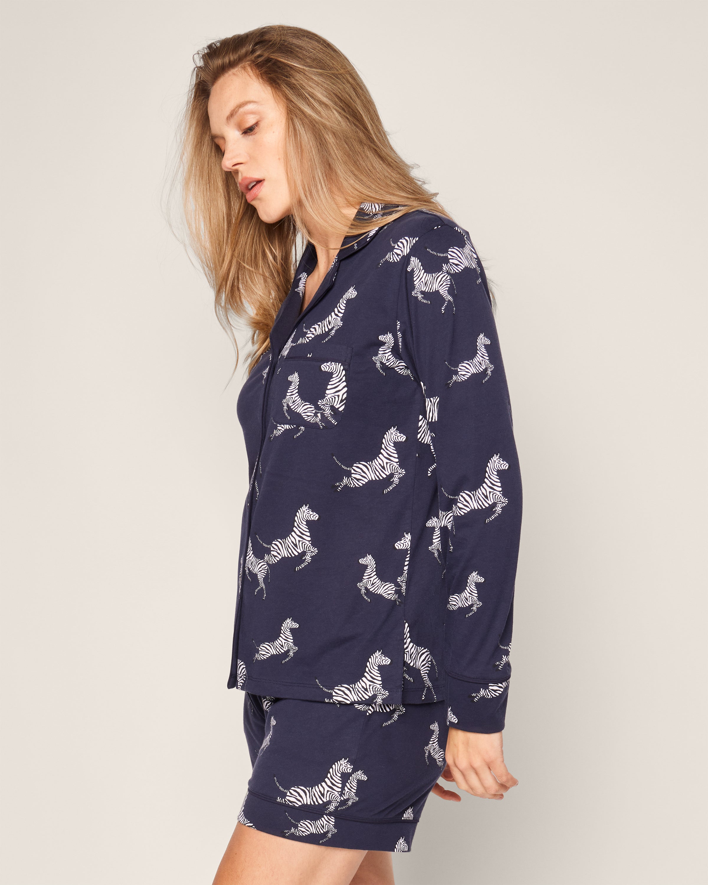 A person with long hair wears the Womens Pima Long Sleeve Short Set in Zèbre de Nuit by Petite Plume, a luxurious Peruvian Pima cotton pajama set featuring a white zebra print on navy blue, elegantly styled with a long-sleeved top and shorts on a light gray background.