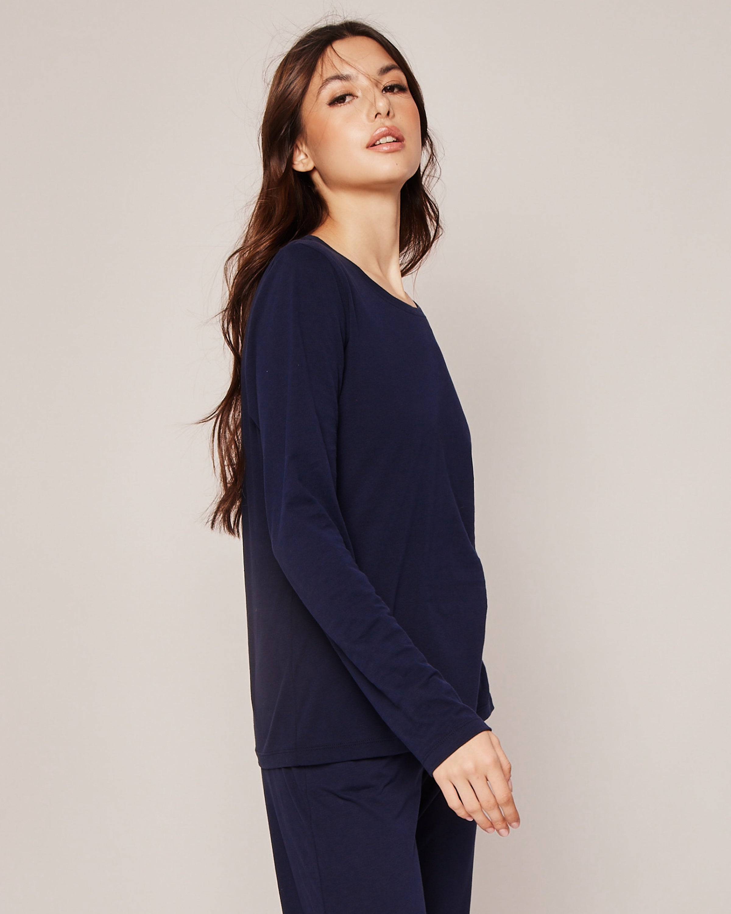 A person with long dark hair is in a navy Womens Pima Long Sleeve Top by Petite Plume and matching pants, crafted from Peruvian Pima cotton. They stand sideways against a plain backdrop, gazing slightly upward with a neutral expression.