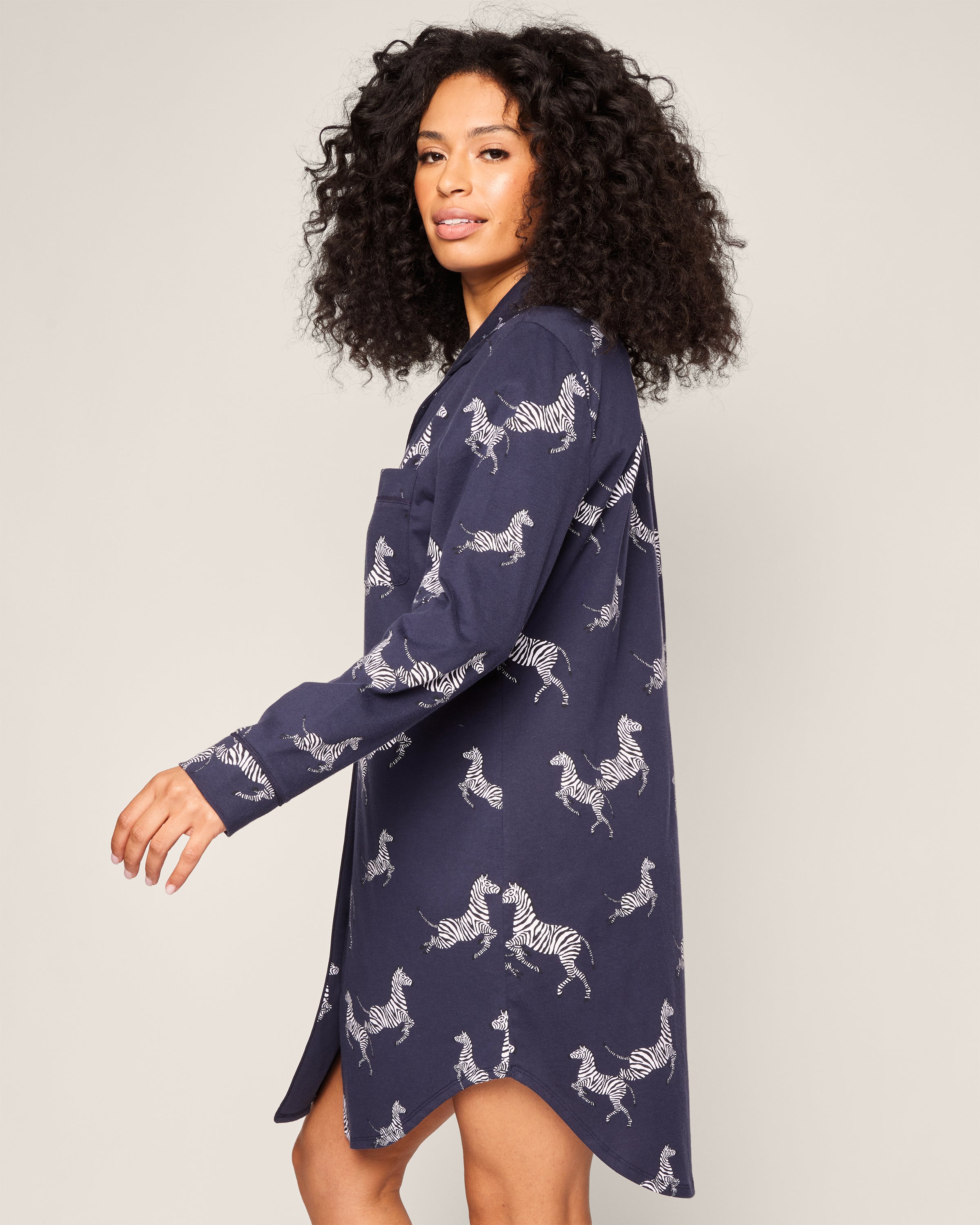 A person with curly hair models the Petite Plume Womens Pima Nightshirt in Zèbre de Nuit, a long navy shirt dress made of soft Peruvian Pima cotton with an eye-catching white zebra print. They stand against a plain background, facing sideways with effortless charm.