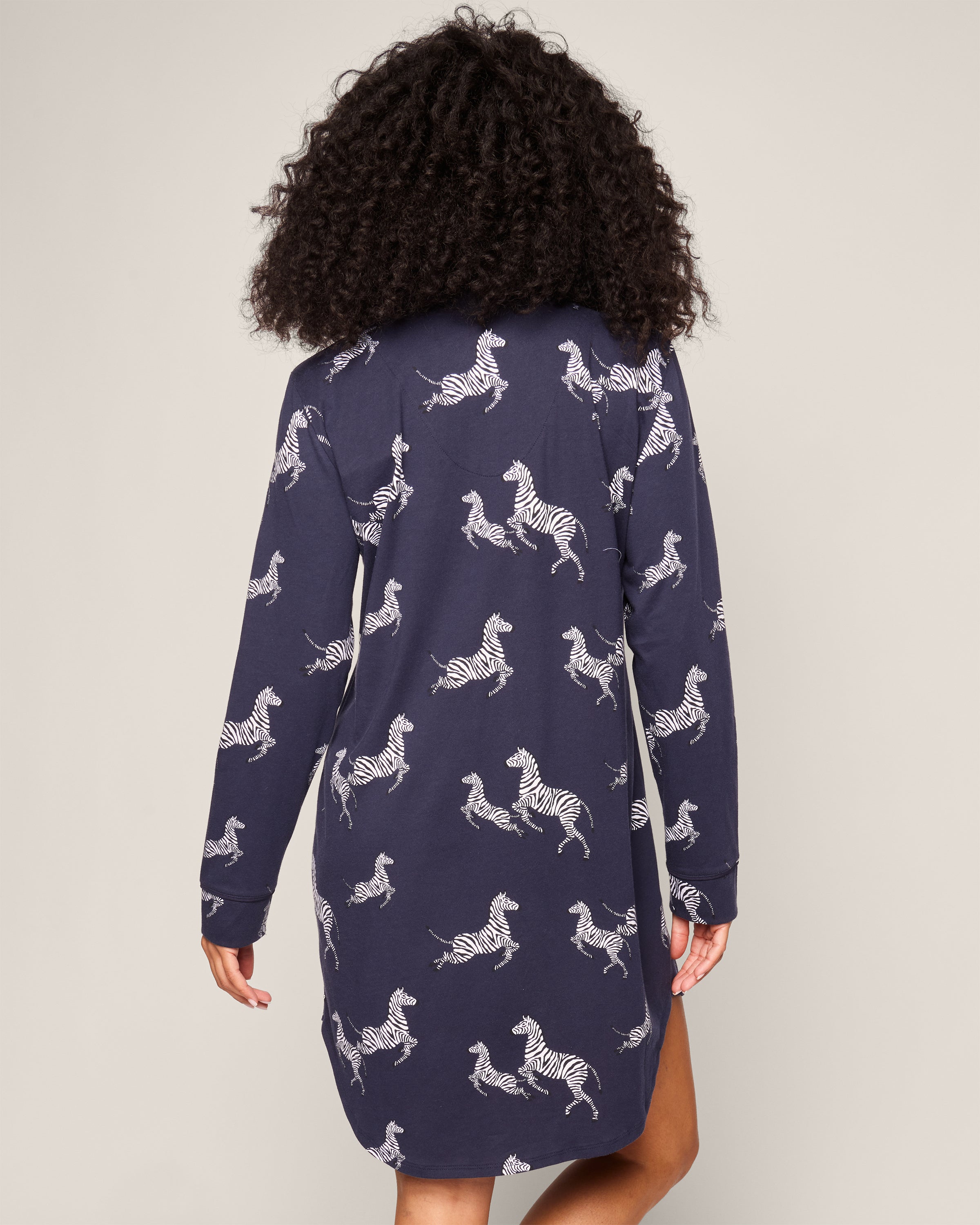 A person with curly hair wears Petite Plumes Womens Pima Nightshirt in Zèbre de Nuit, a navy blue zebra print with long sleeves, falling just above the knees. The background is a plain light color.