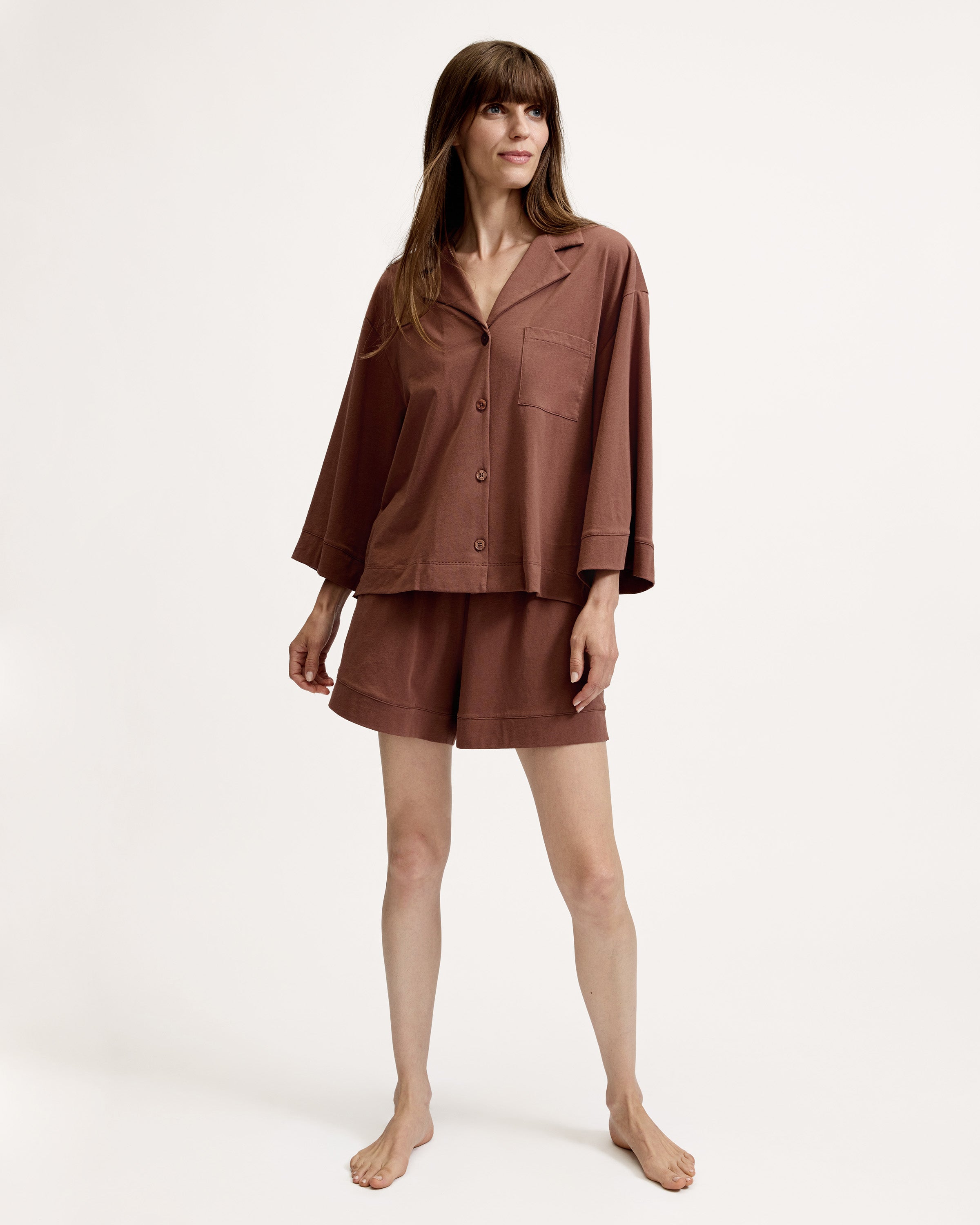A woman stands barefoot against a white background, wearing The Olivia Lightweight Pima Short Set in Chocolate Brown—featuring a loose-fitting, long-sleeved button-up top with a pocket and matching shorts.