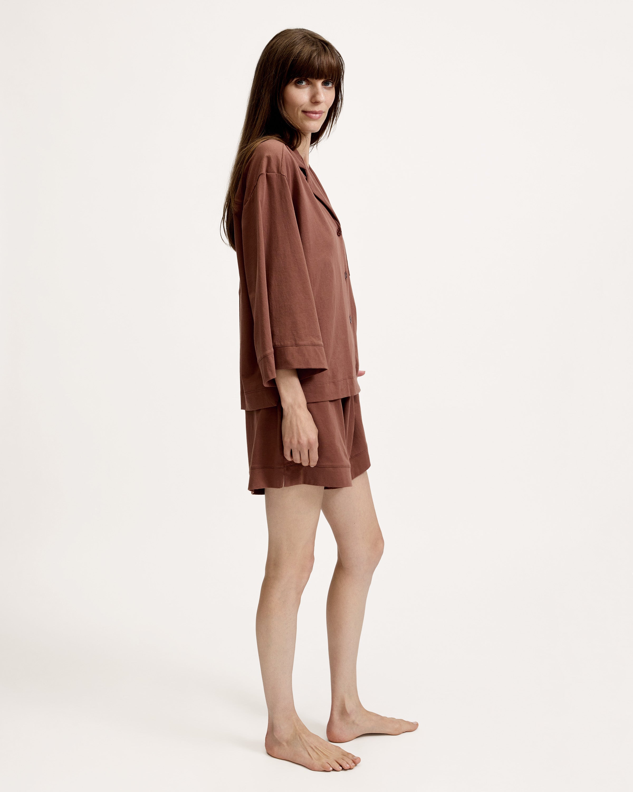 A woman stands barefoot against a white background, wearing The Olivia Lightweight Pima Short Set in Chocolate Brown—featuring a loose-fitting, long-sleeved button-up top with a pocket and matching shorts.