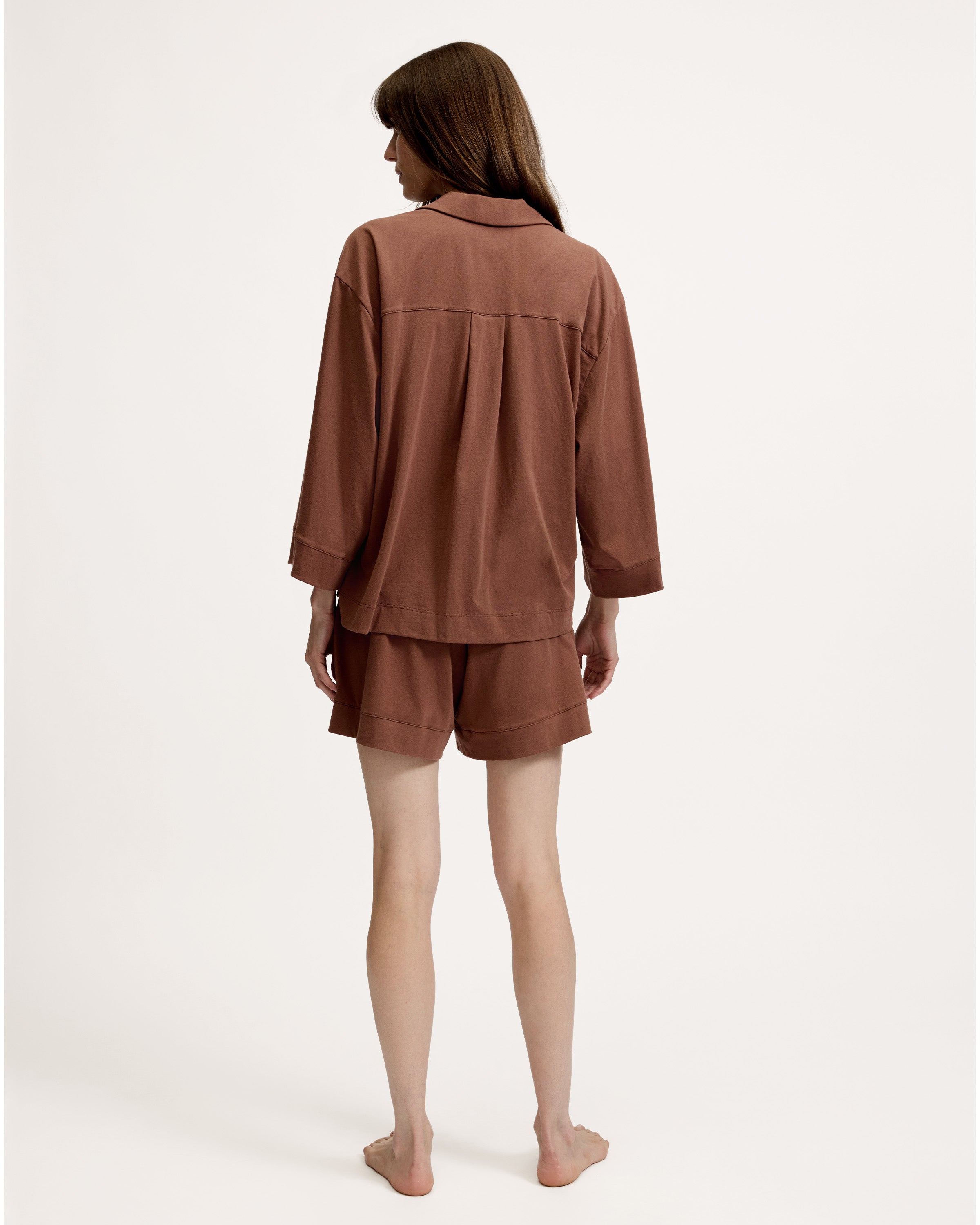 A woman stands barefoot against a white background, wearing The Olivia Lightweight Pima Short Set in Chocolate Brown—featuring a loose-fitting, long-sleeved button-up top with a pocket and matching shorts.