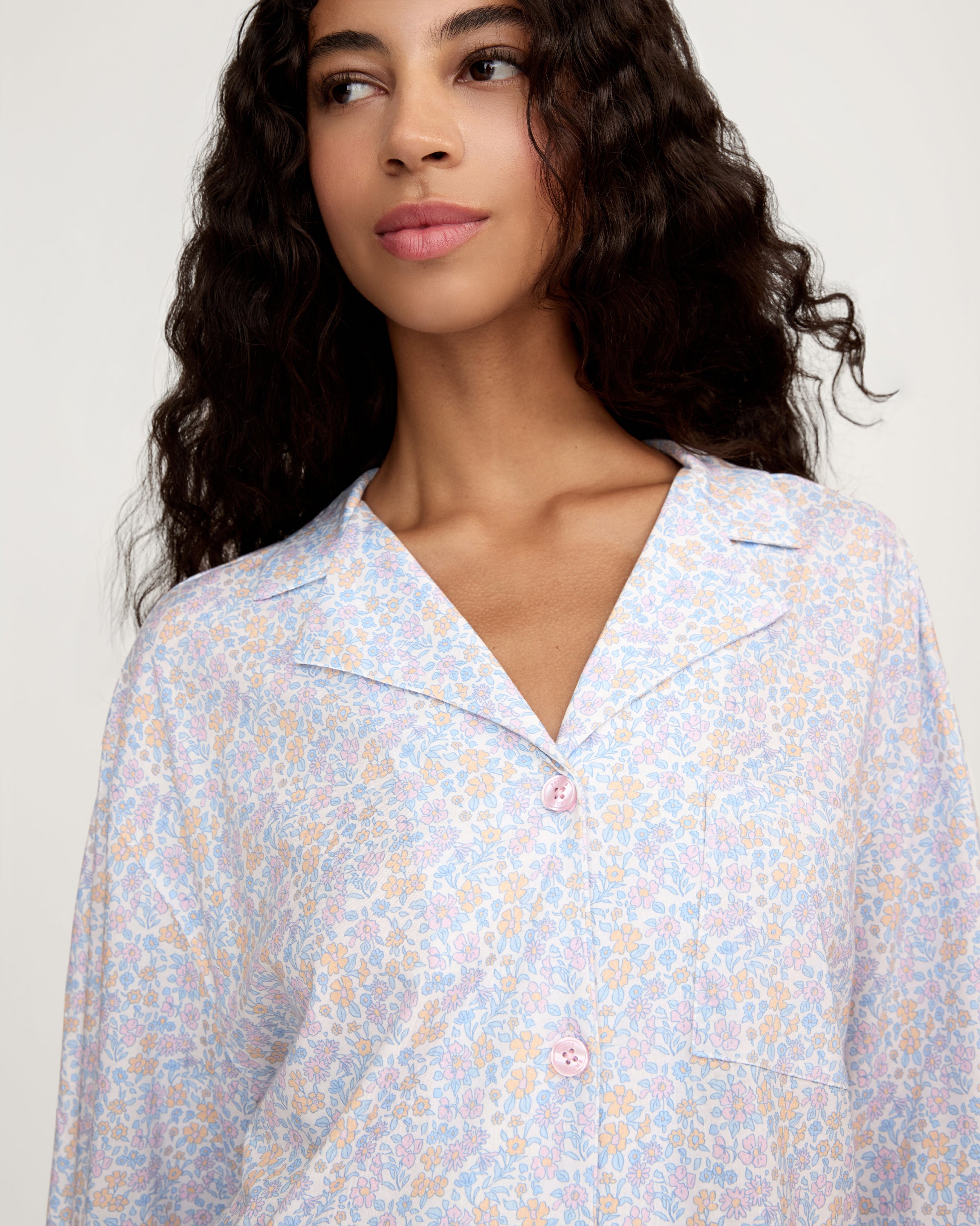 A woman with long, wavy hair wears The Olivia Lightweight Pima Short Set in Fleur Blanche, featuring a loose button-up top with wide sleeves and matching shorts adorned with a subtle floral pattern. She stands against a plain, light background.
