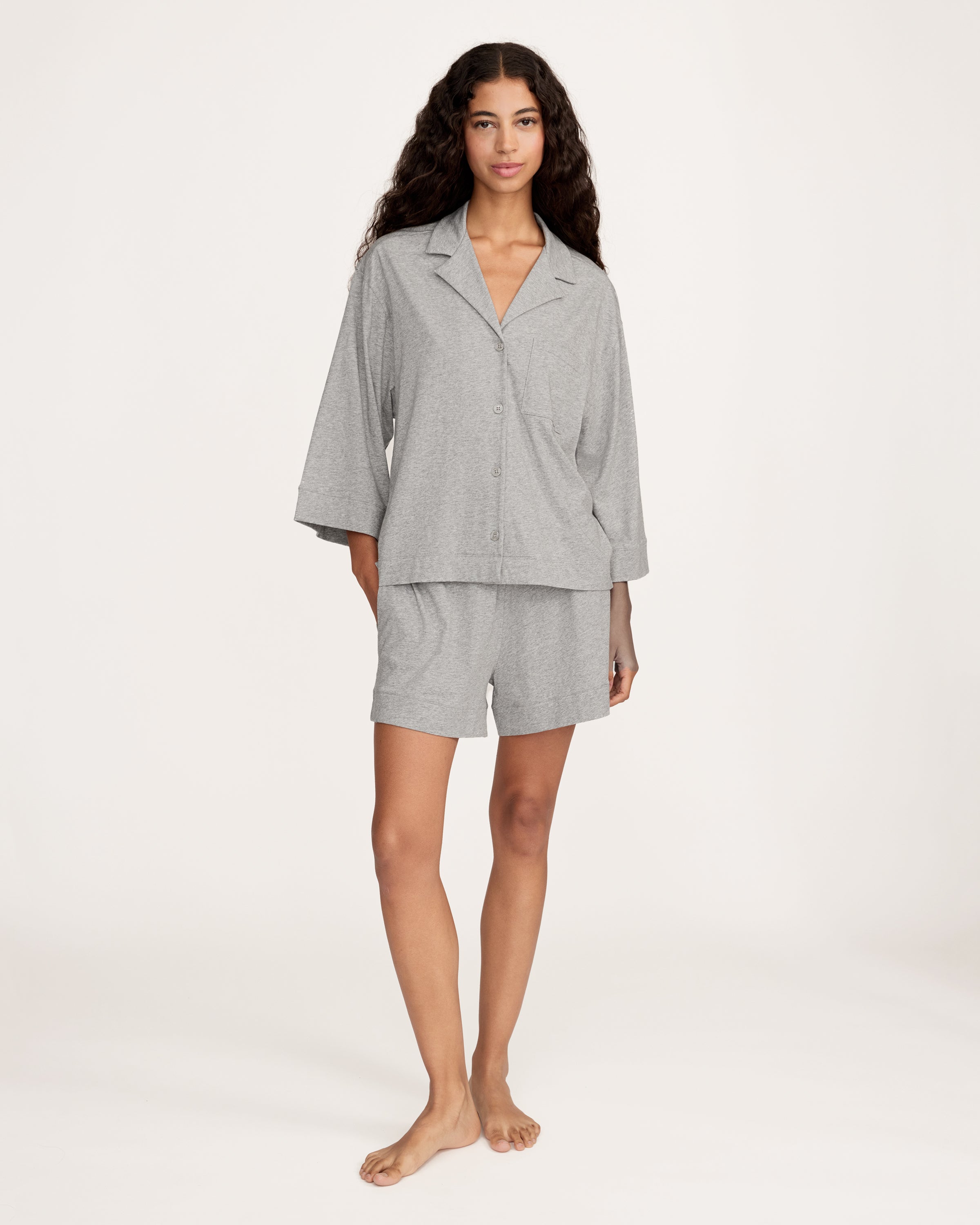A woman with long, wavy hair and a neutral expression stands barefoot against a plain background, wearing The Olivia Lightweight Pima Short Set in Heather Grey—featuring a loose-fitting, long-sleeve top and matching shorts.