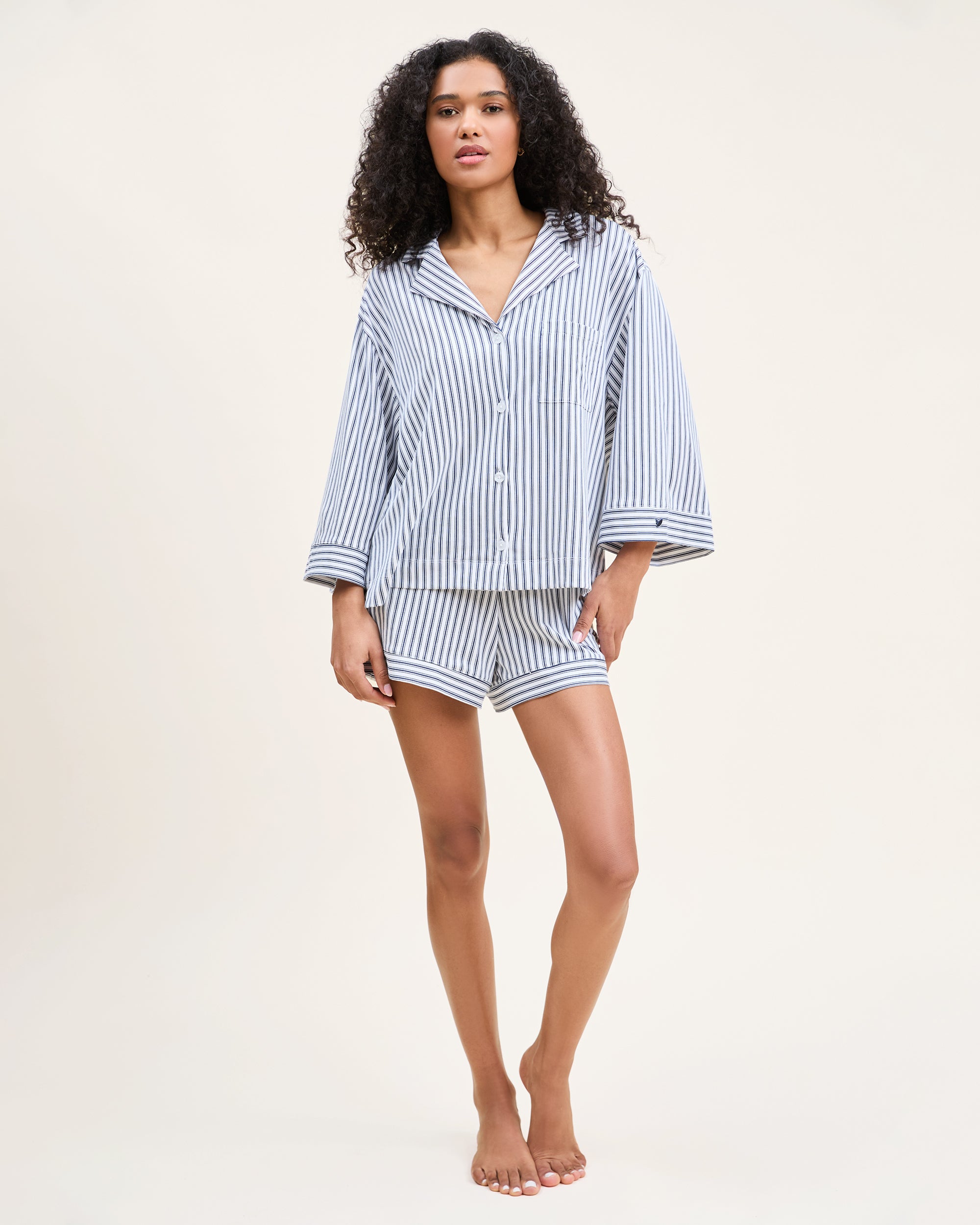 The Olivia Pima Short Set in Navy French Ticking features a matching set of blue and white vertical striped pajamas with a long-sleeve, button-up shirt with chest pocket and drawstring shorts.