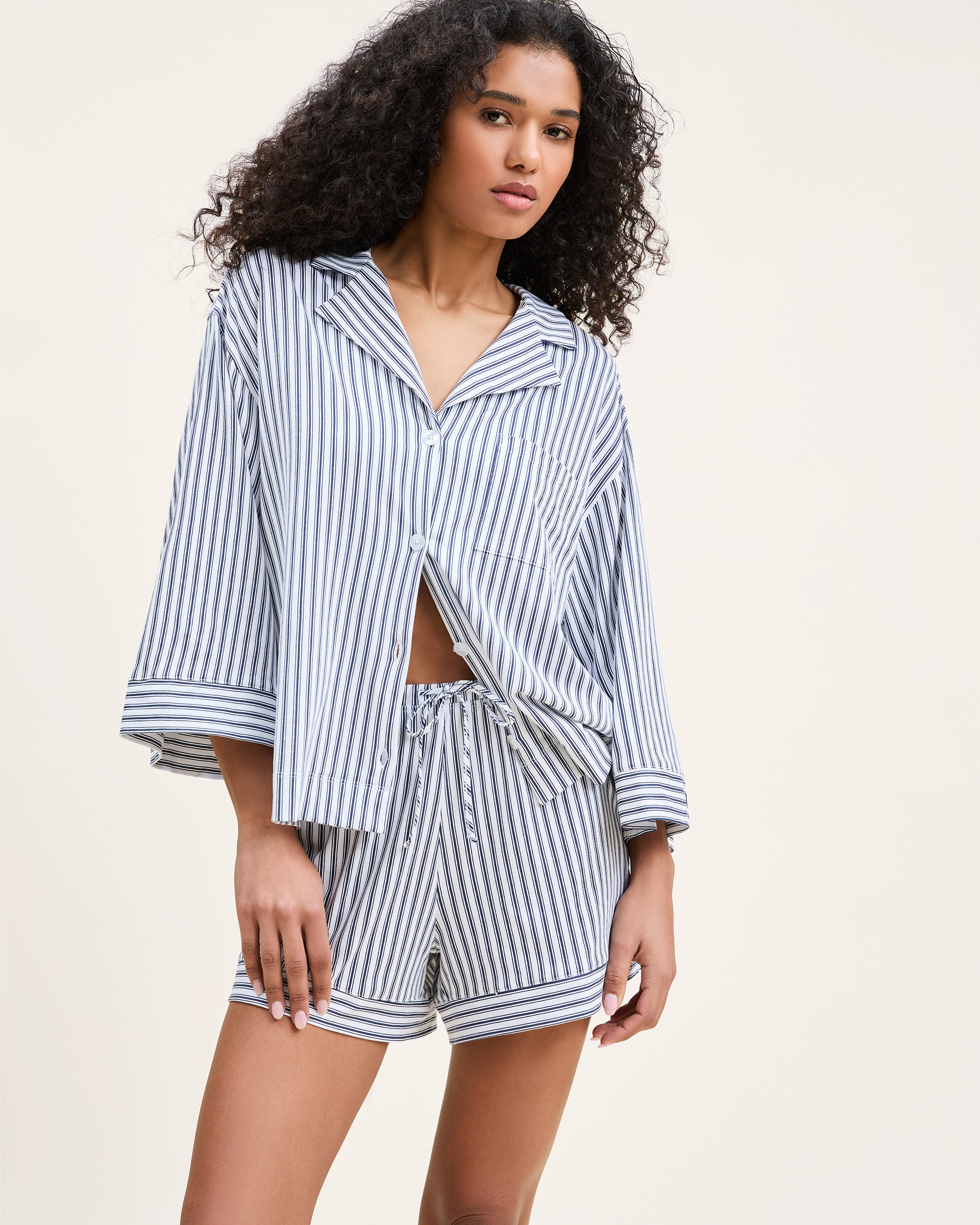 The Olivia Pima Short Set in Navy French Ticking features a matching set of blue and white vertical striped pajamas with a long-sleeve, button-up shirt with chest pocket and drawstring shorts.