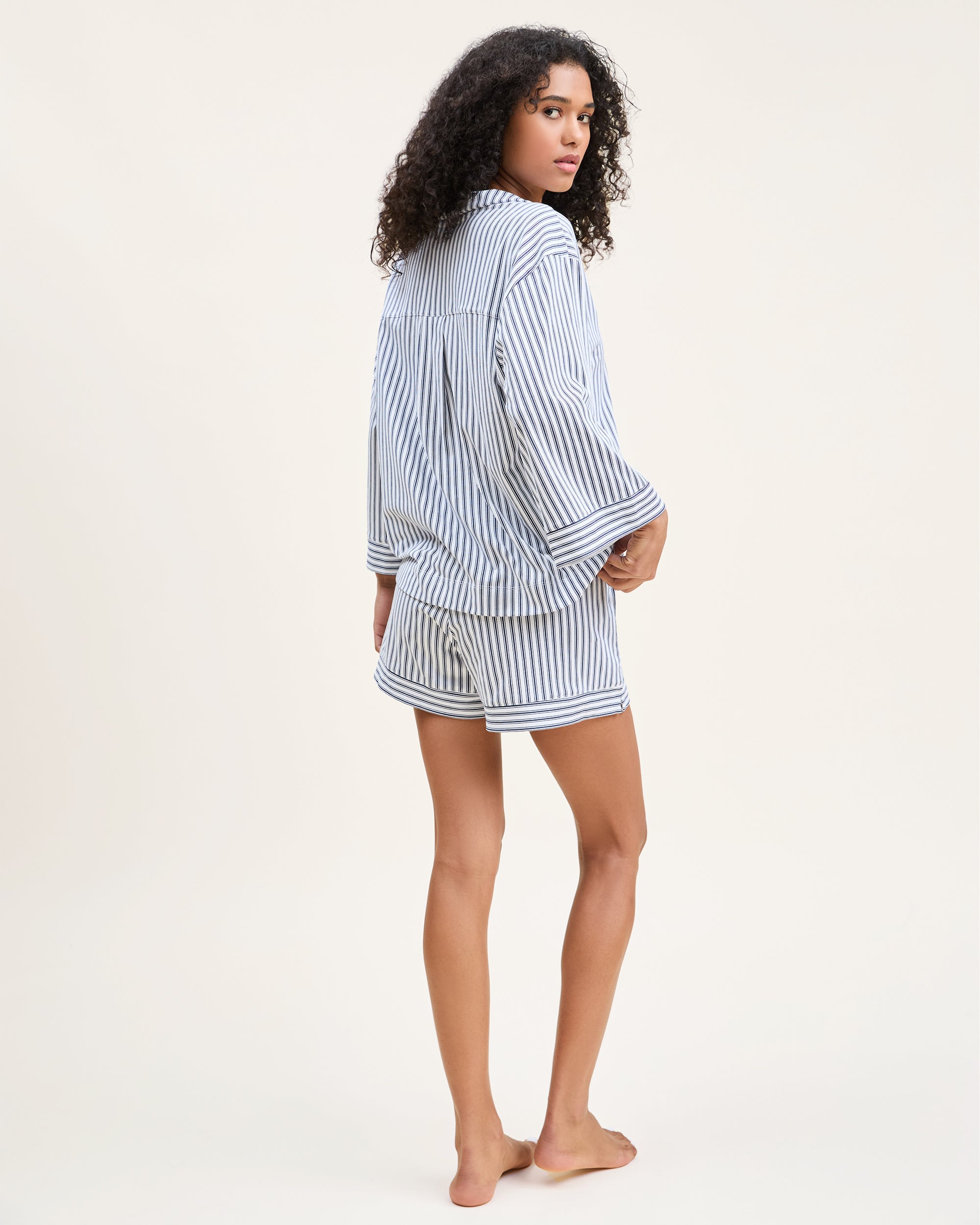 The Olivia Pima Short Set in Navy French Ticking features a matching set of blue and white vertical striped pajamas with a long-sleeve, button-up shirt with chest pocket and drawstring shorts.