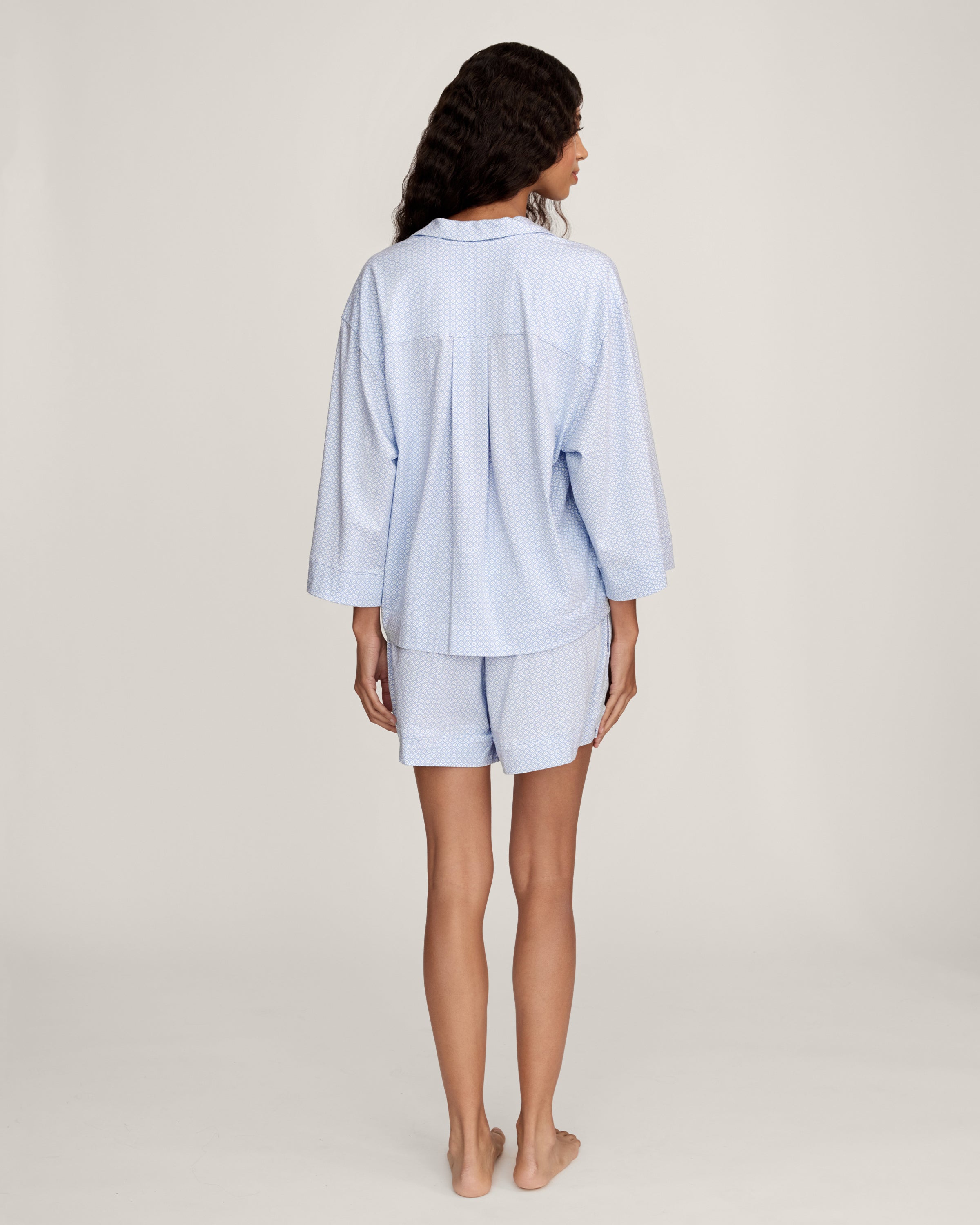 A woman with long curly hair models The Olivia Lightweight Pima Short Set in Periwinkle Geo—a light blue, long-sleeved button-up pajama top and matching dotted shorts—against a plain light background.