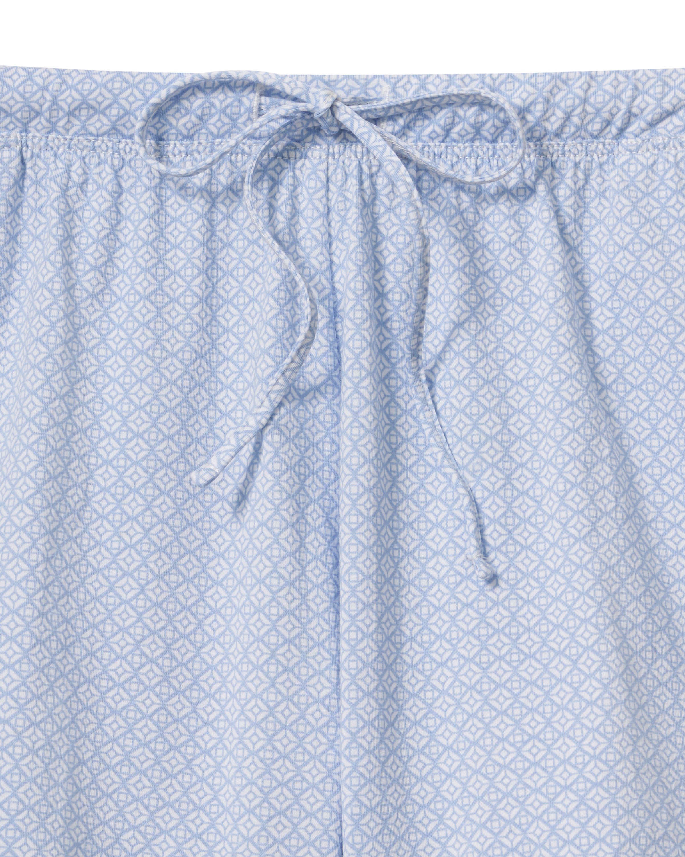 A woman with long curly hair models The Olivia Lightweight Pima Short Set in Periwinkle Geo—a light blue, long-sleeved button-up pajama top and matching dotted shorts—against a plain light background.