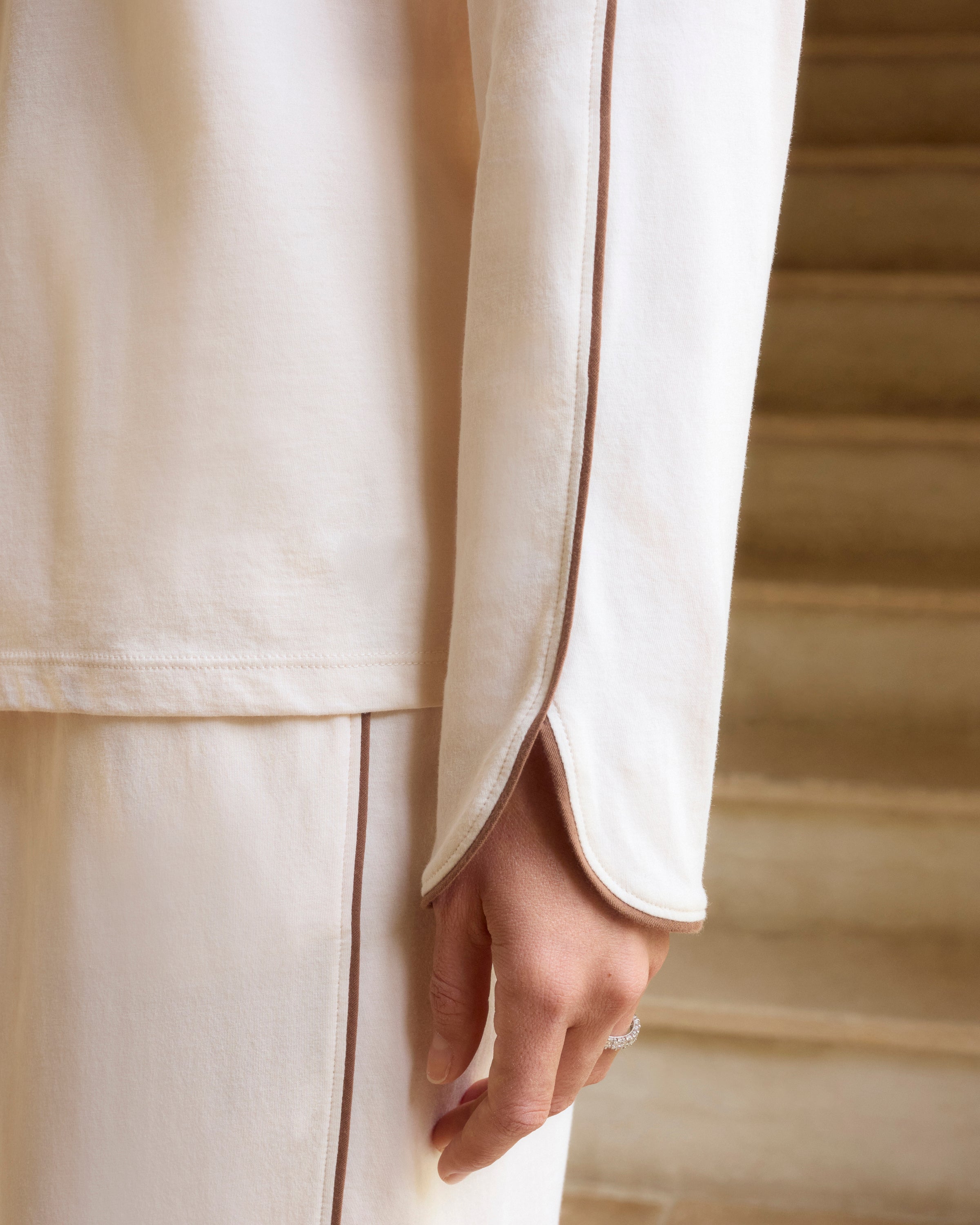 A person stands barefoot on a white background, wearing The Margaux Lightweight Pima Pajama Set in Ivory, featuring long sleeves, buttons, and pants with dark piping along the edges.