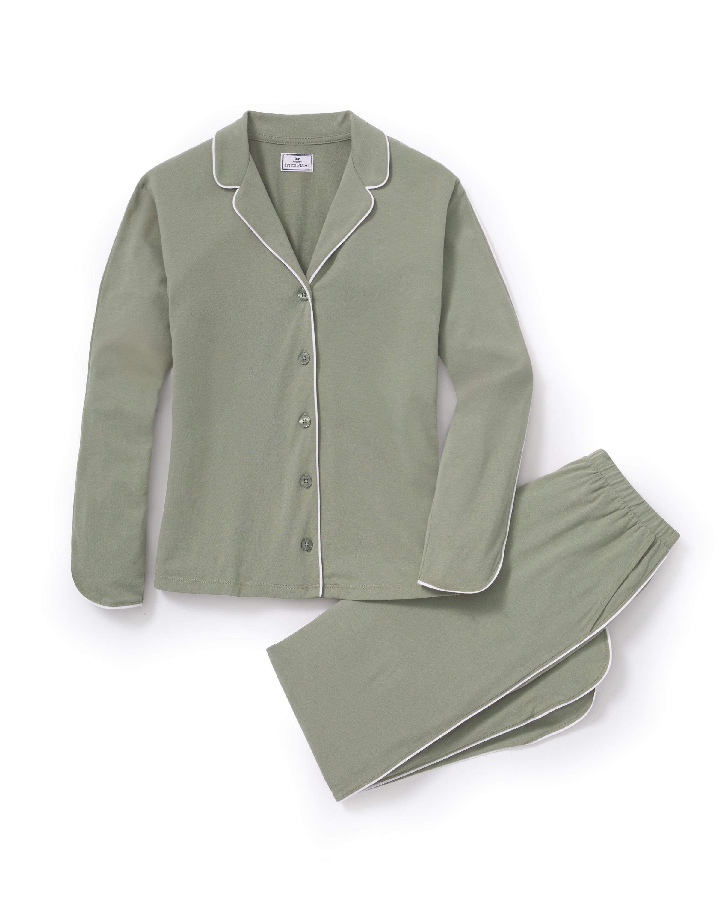 A woman with long dark hair wears The Margaux Pima Pajama Set in Sage, featuring a button-up long-sleeve top with white piping and matching pants. She stands barefoot against a plain, light background.