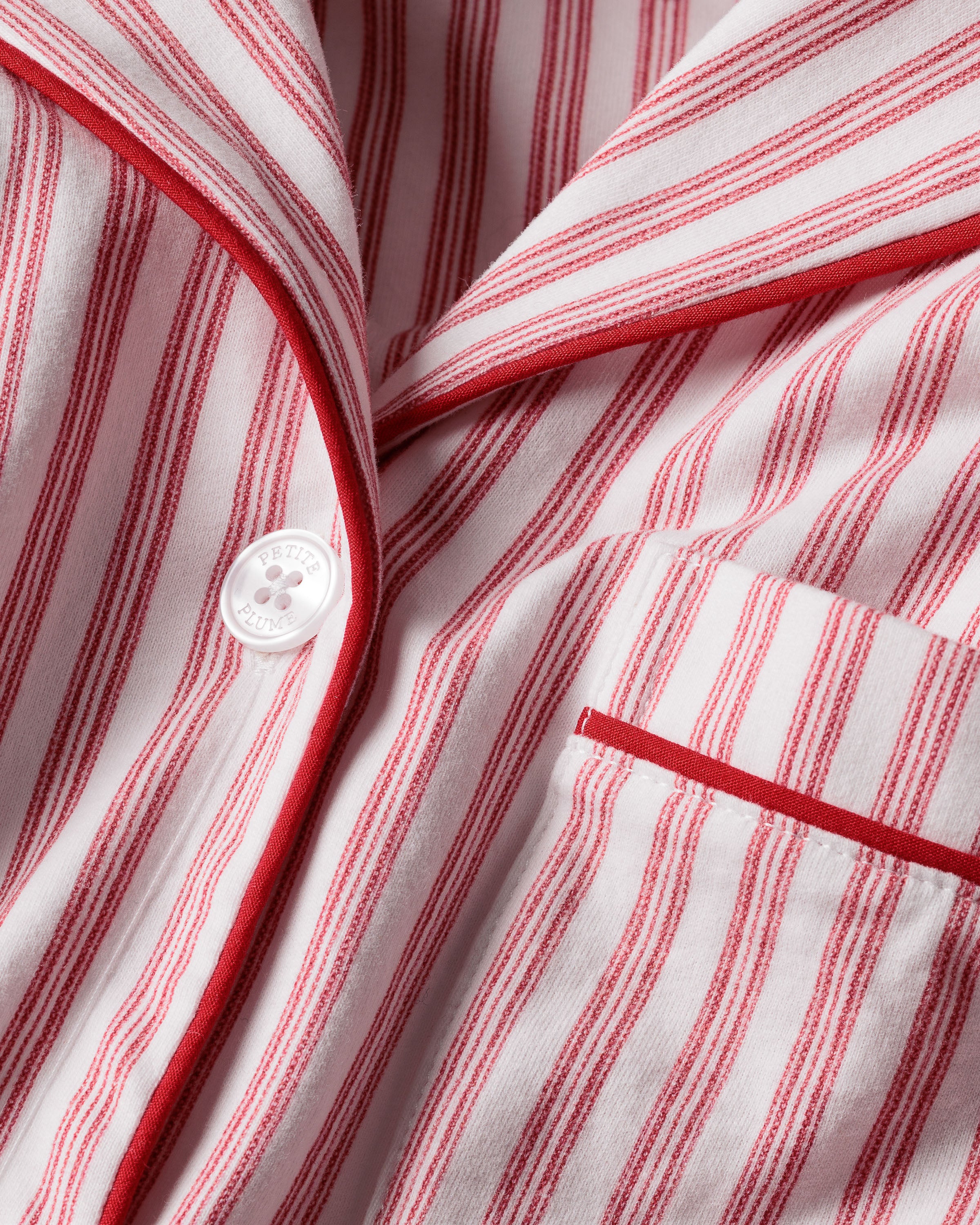Close-up of Petite Plumes Womens Pima Pajama Set in Antique Red Ticking, showcasing luxury fabric with red and white vertical stripes. The image reveals part of the garment with a white button, collar, and pocket trimmed in red, hinting at its soft and cozy Peruvian Pima cotton.