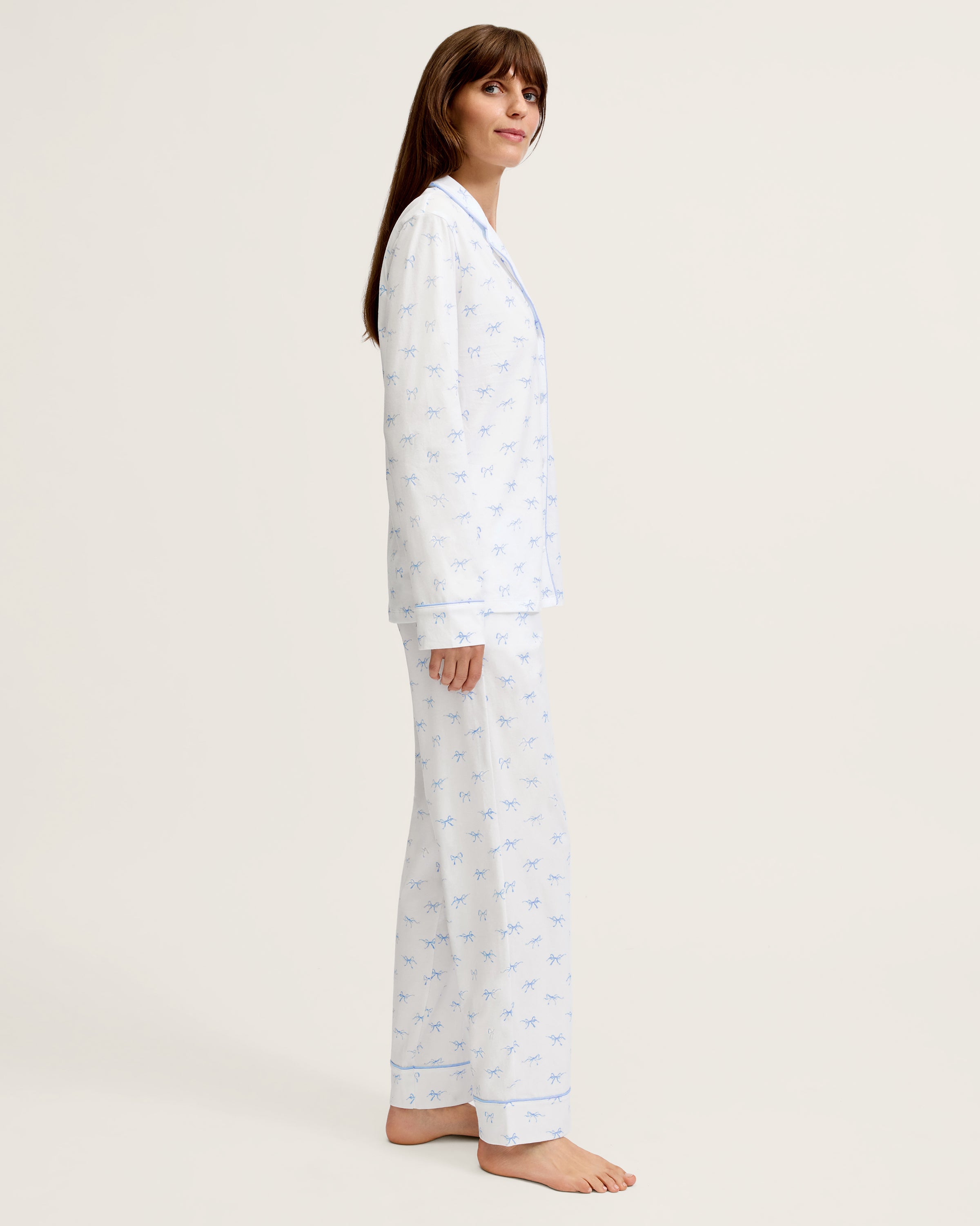 The Women's Pima Pajama Set in Something Blue Bows is a white set with blue piping, including a collared button-up top with chest pocket and matching pants. Both feature a repeating blue print of small bows.