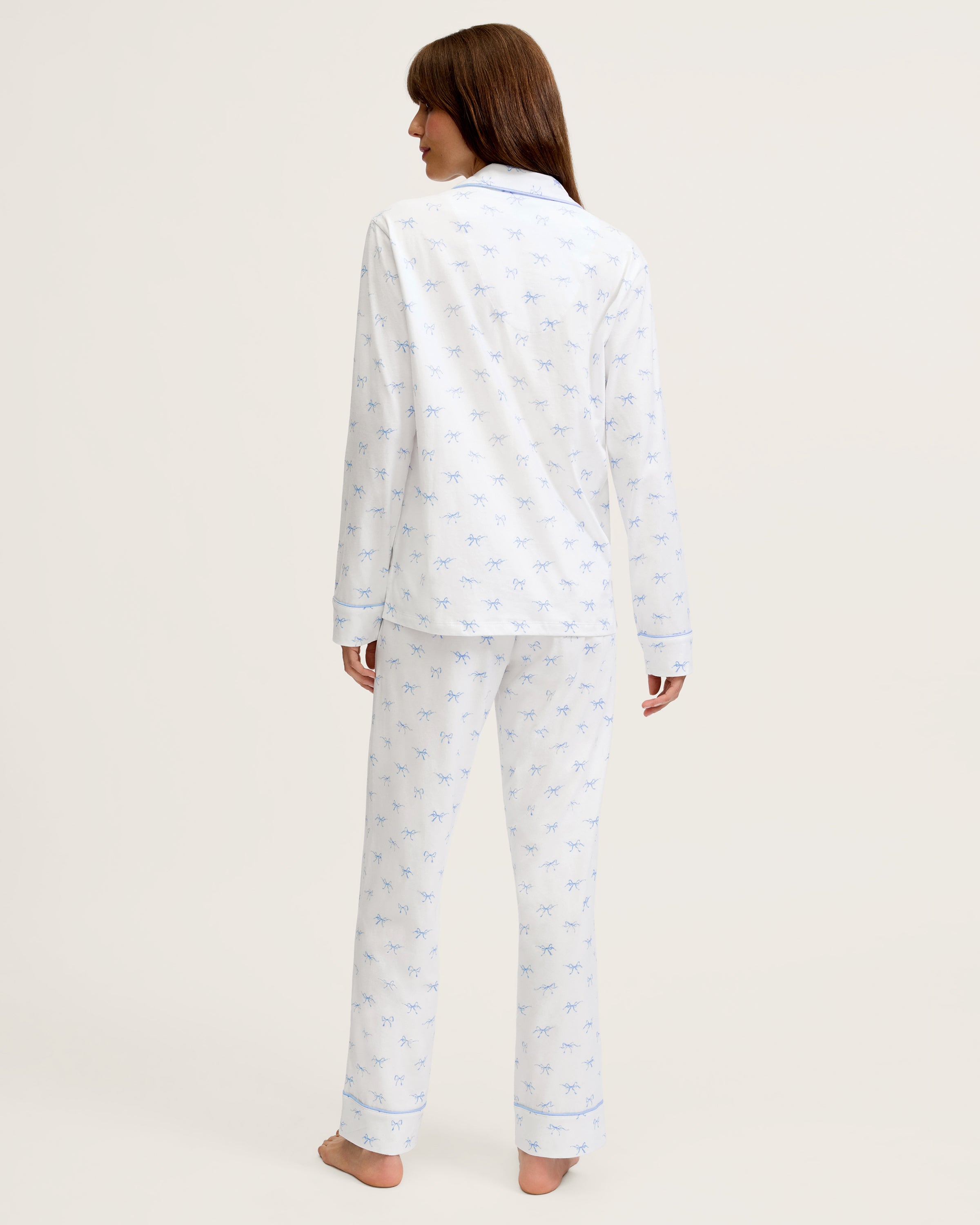 The Women's Pima Pajama Set in Something Blue Bows is a white set with blue piping, including a collared button-up top with chest pocket and matching pants. Both feature a repeating blue print of small bows.