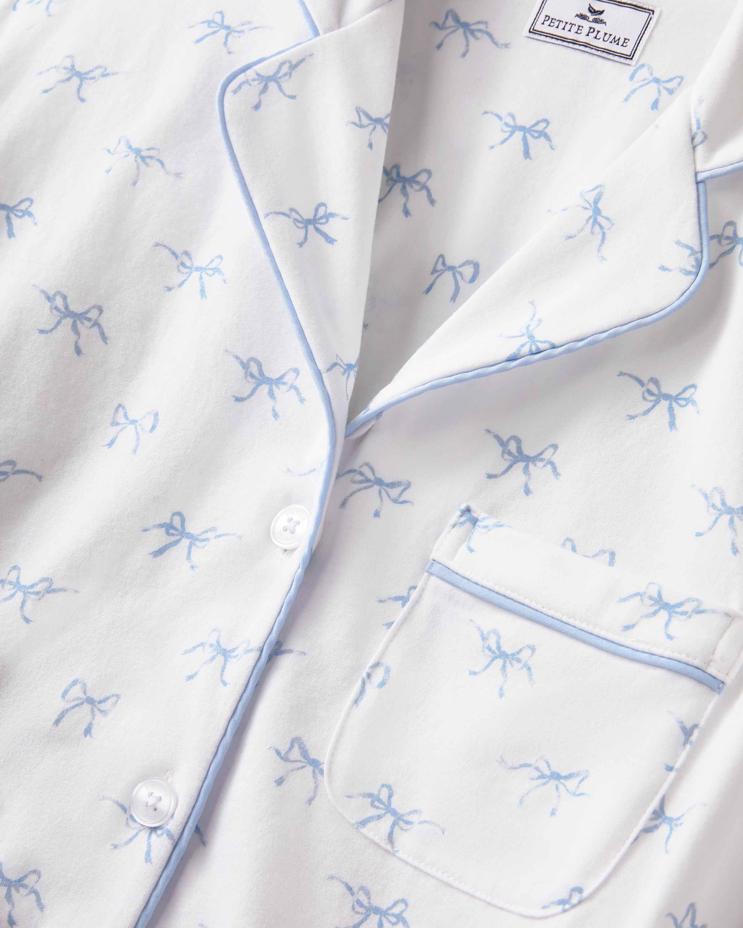 The Women's Pima Pajama Set in Something Blue Bows is a white set with blue piping, including a collared button-up top with chest pocket and matching pants. Both feature a repeating blue print of small bows.
