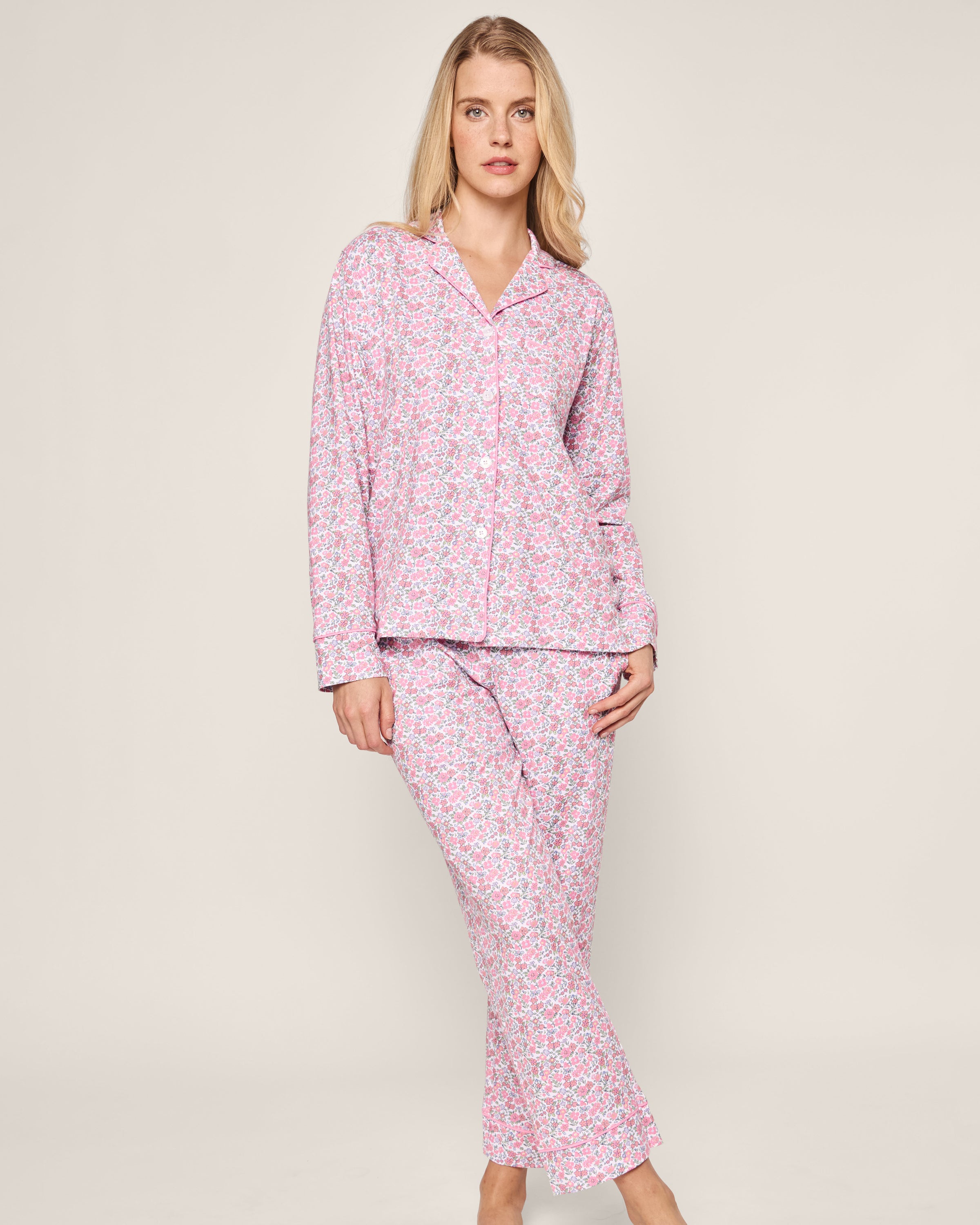 A person wears the Womens Pima Pajama Set in Fleurs de Rose by Petite Plume, featuring a long-sleeved, collared top. They stand barefoot against a plain light-colored background.