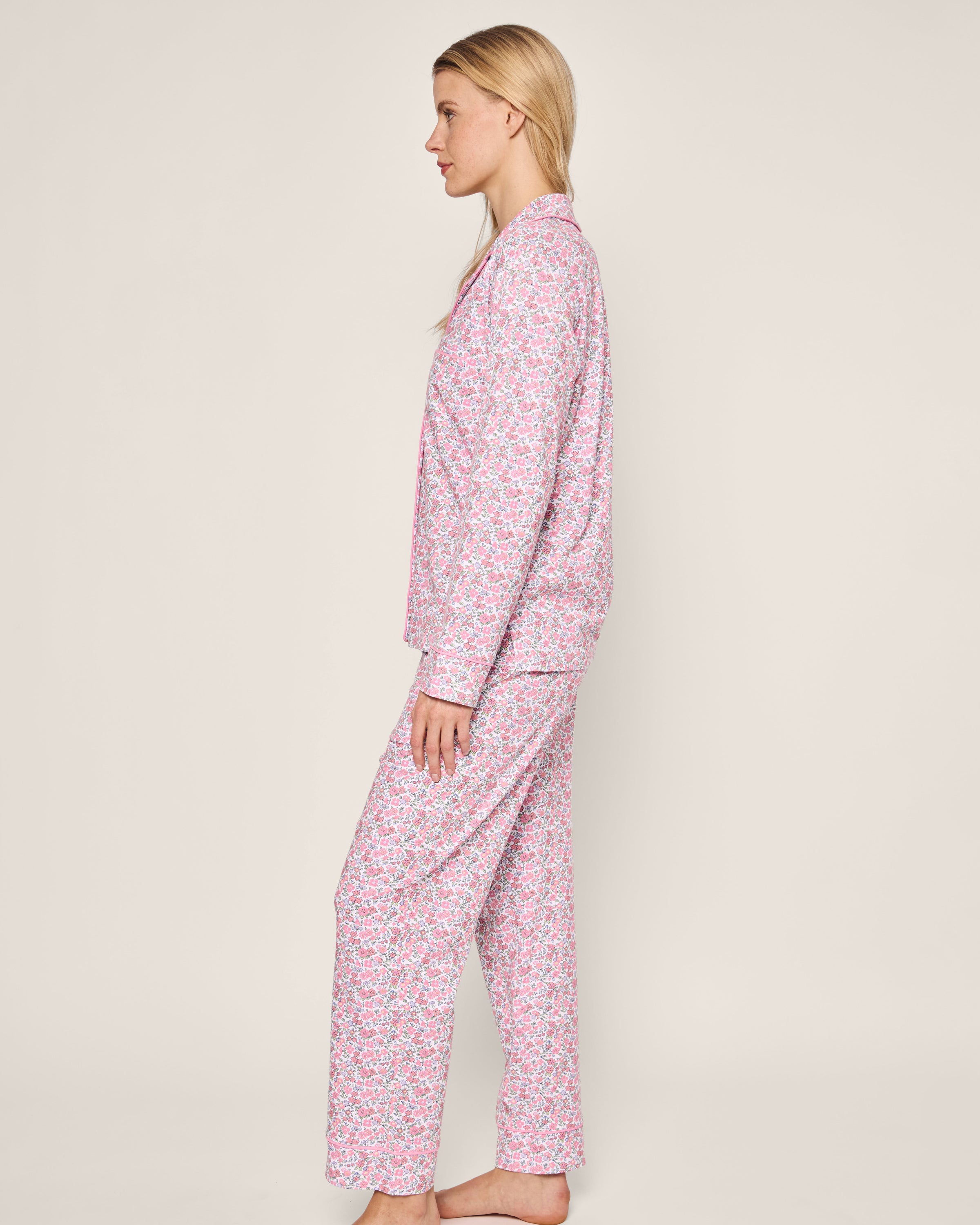 A person with long blonde hair stands sideways, modeling the luxurious Womens Pima Pajama Set in Fleurs de Rose by Petite Plume. The plain, light-colored background enhances the elegant floral print of this exquisite sleepwear.