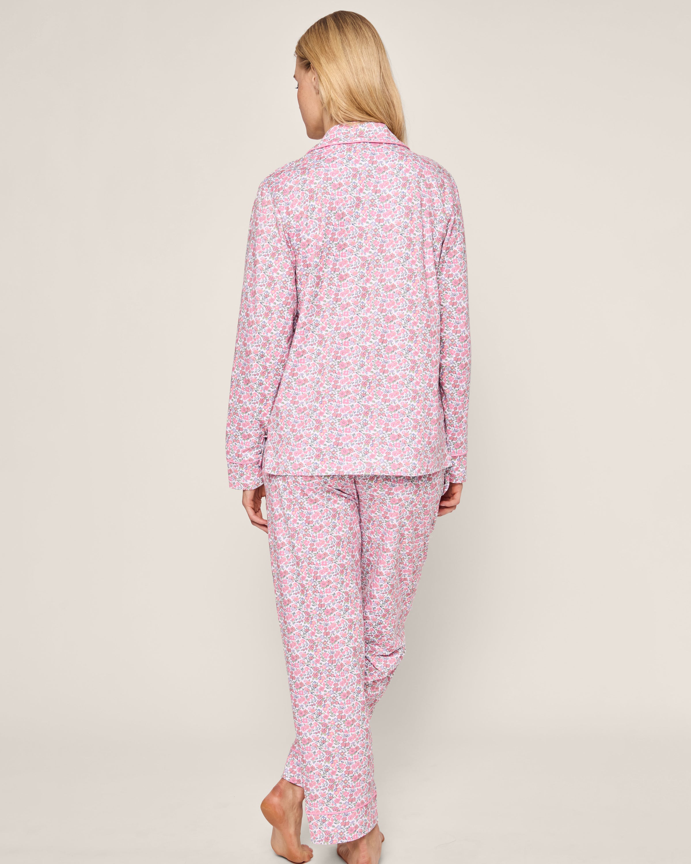 A person with long blonde hair, seen from the back, wears a Womens Pima Pajama Set in Fleurs de Rose by Petite Plume. The luxury sleepwear features a long-sleeved top and pants with a pink floral design. They stand barefoot against a plain, neutral background.