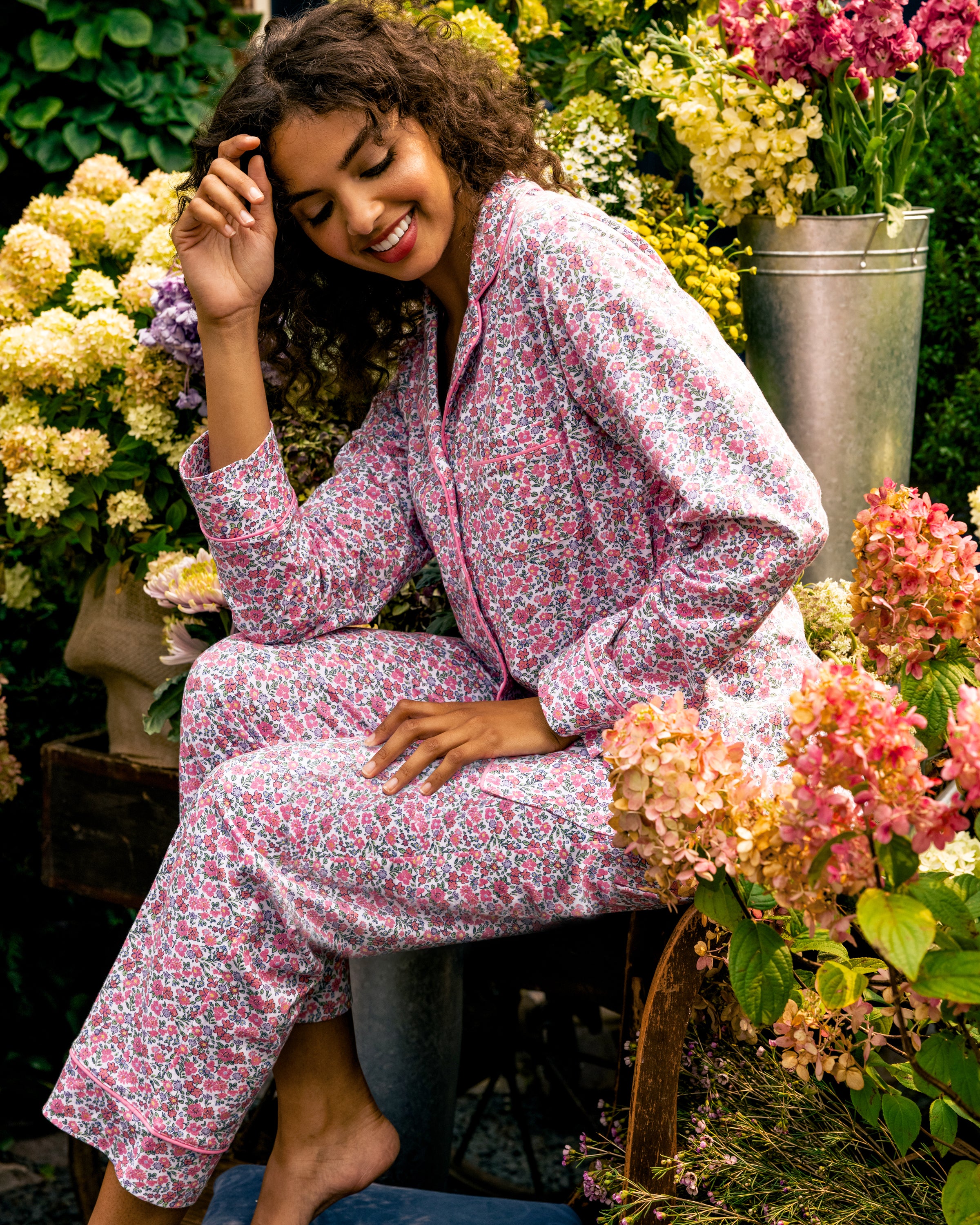 Wearing a Womens Pima Pajama Set in Fleurs de Rose by Petite Plume, a person sits in a garden amidst colorful hydrangeas, smiling with one hand on their head, embodying the relaxed luxury of their sleepwear.