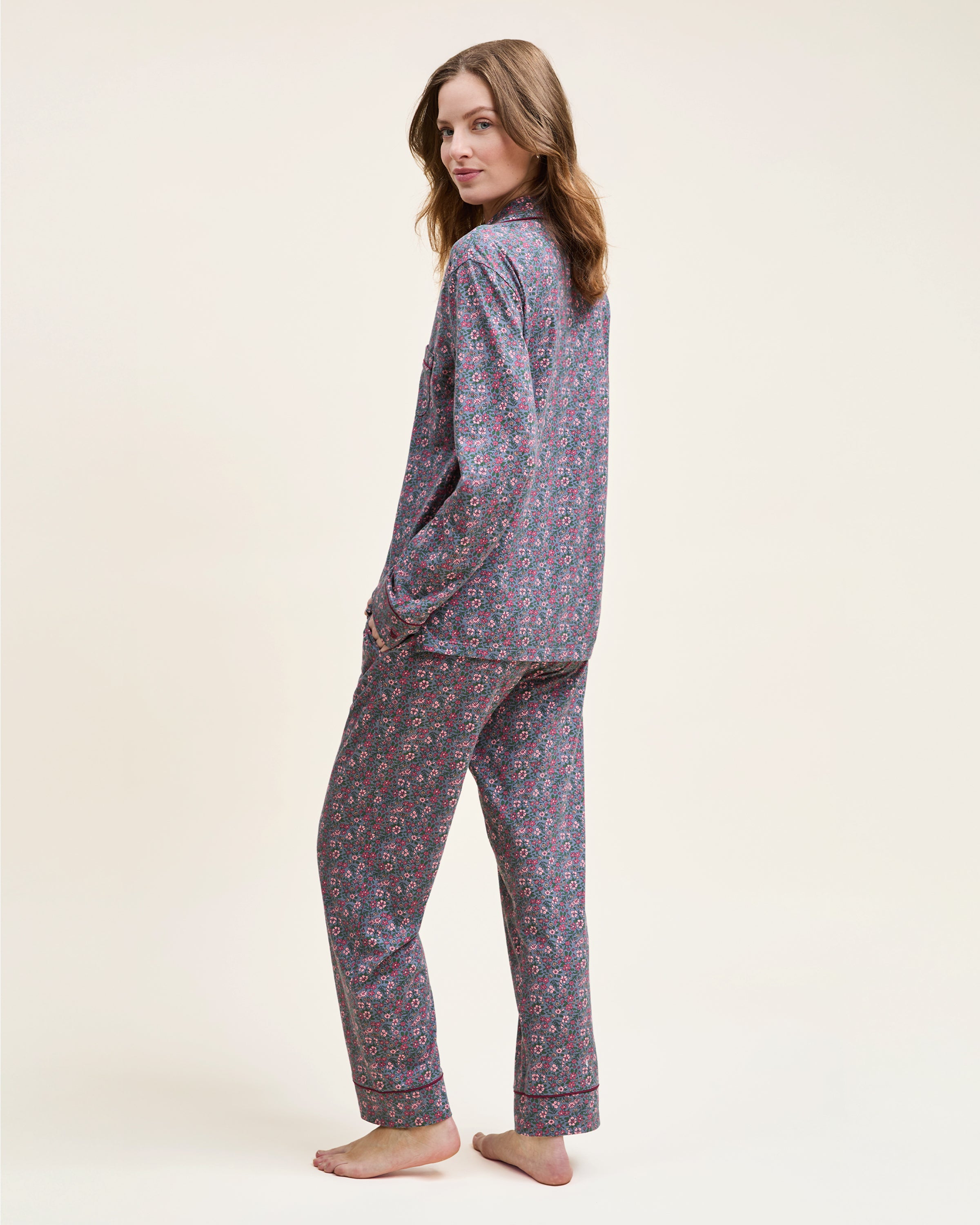 The Women's Pima Pajama Set in Fleurs de Saison features long sleeves and a small floral print in pink, white, and blue on gray. The set includes a button-up top with a chest pocket and matching pants.