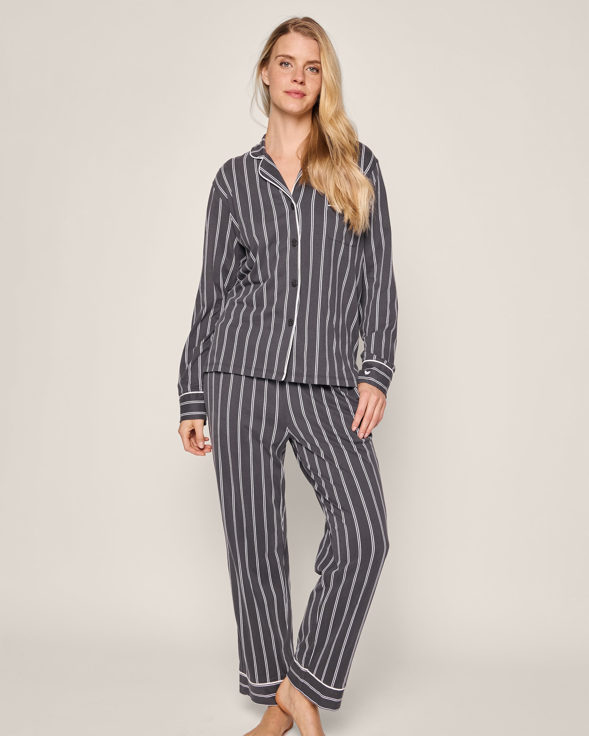 A woman with long blonde hair is wearing Petite Plumes Womens Pima Pajama Set in Grey Stripe. This luxe sleepwear features a dark base with light stripes, crafted from Pima cotton. She stands barefoot and relaxed against a plain backdrop.
