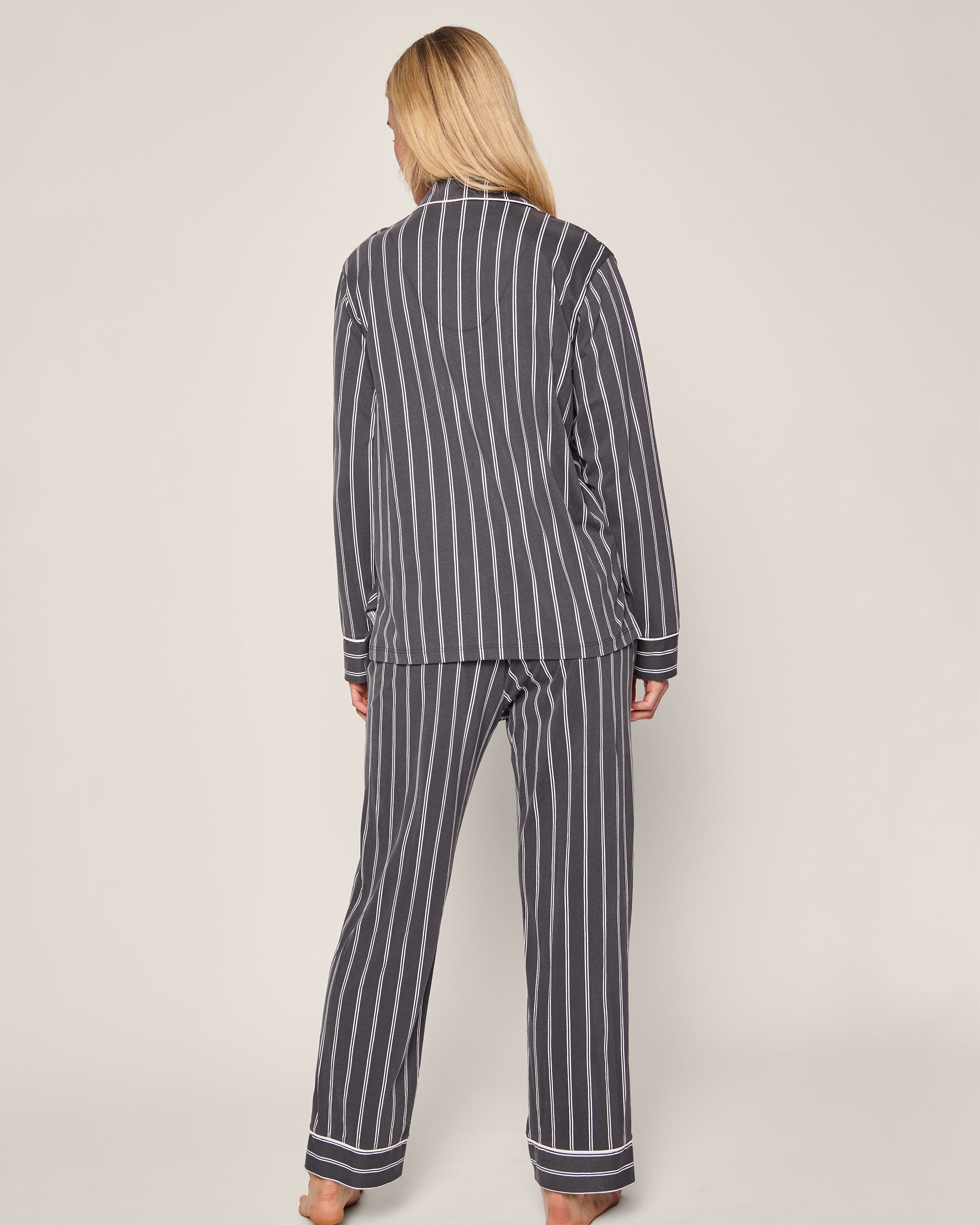 A person with long blond hair wears the luxurious Petite Plume Womens Pima Pajama Set in Grey Stripe. Made from soft Pima cotton, these timeless pajamas provide ultimate comfort as they stand barefoot on a simple, light-colored background.