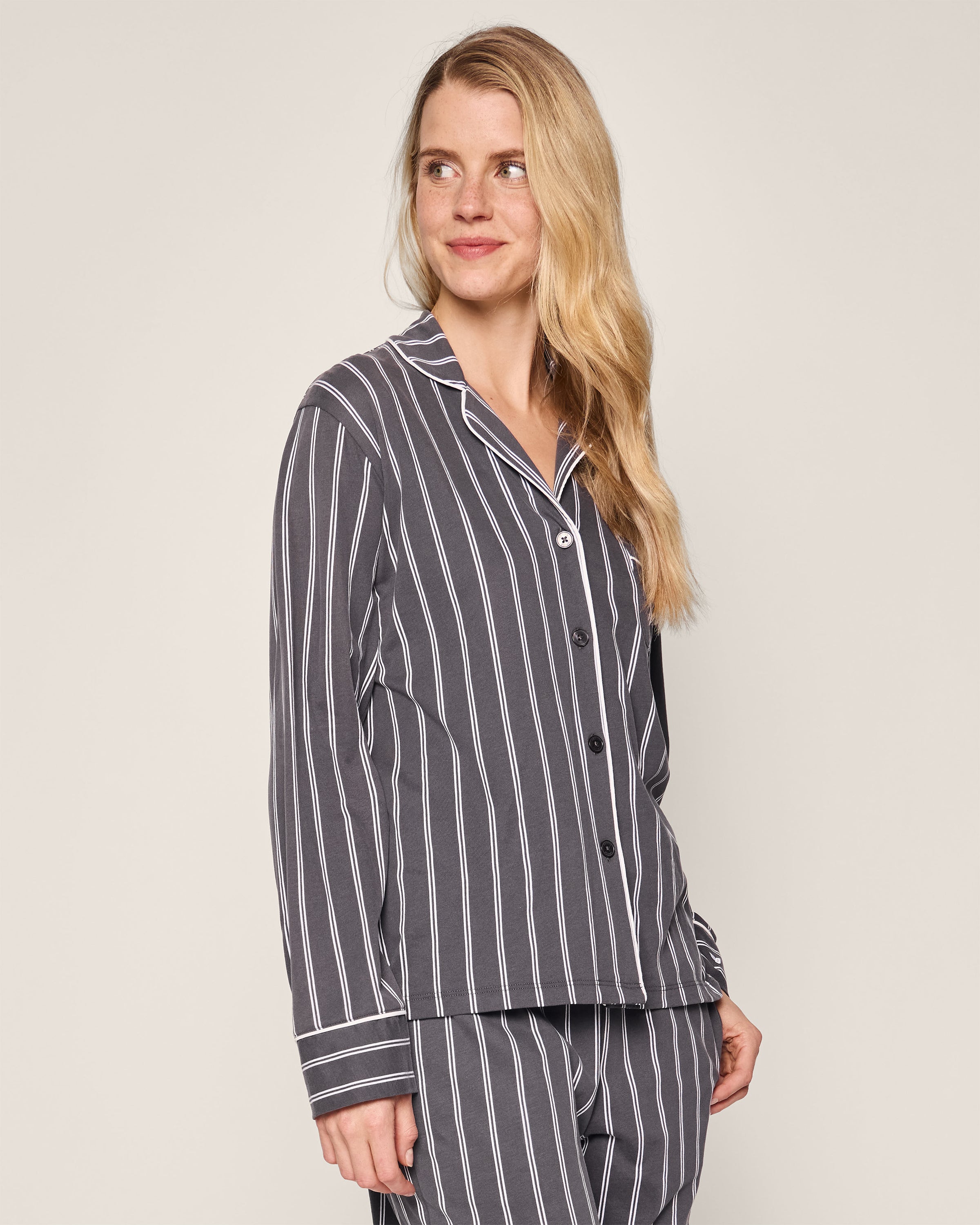 A person with long blonde hair wears the Womens Pima Pajama Set in Grey Stripe from Petite Plume. The elegant top has a collar and buttons. Smiling, the individual stands against a plain, light-colored background.