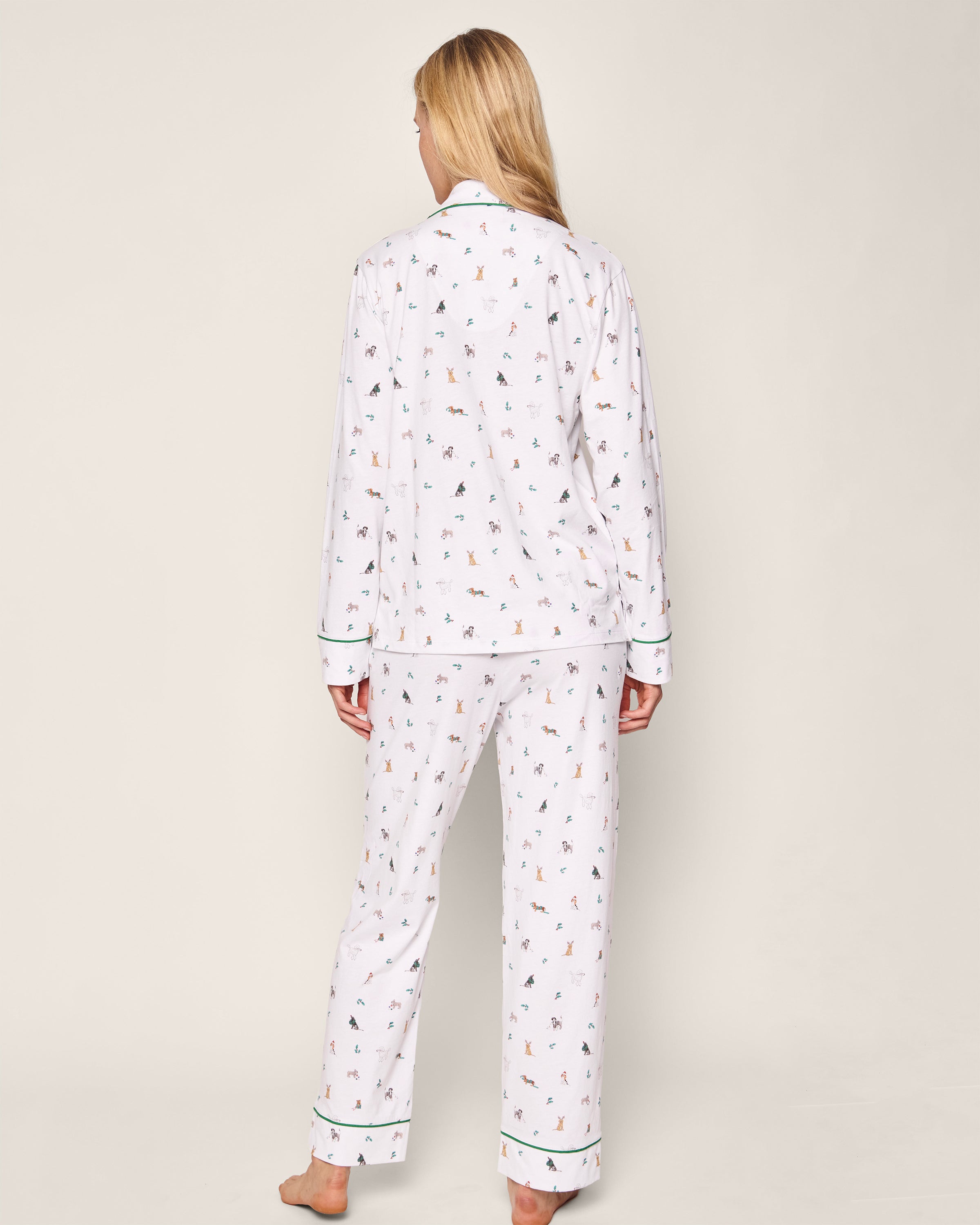 A person with long blonde hair is shown from the back wearing Petite Plumes Womens Pima Pajama Set in Jingle Paws, made of luxurious cotton with small multicolored animal prints and green piping on the sleeves and cuffs, set against a plain off-white background.