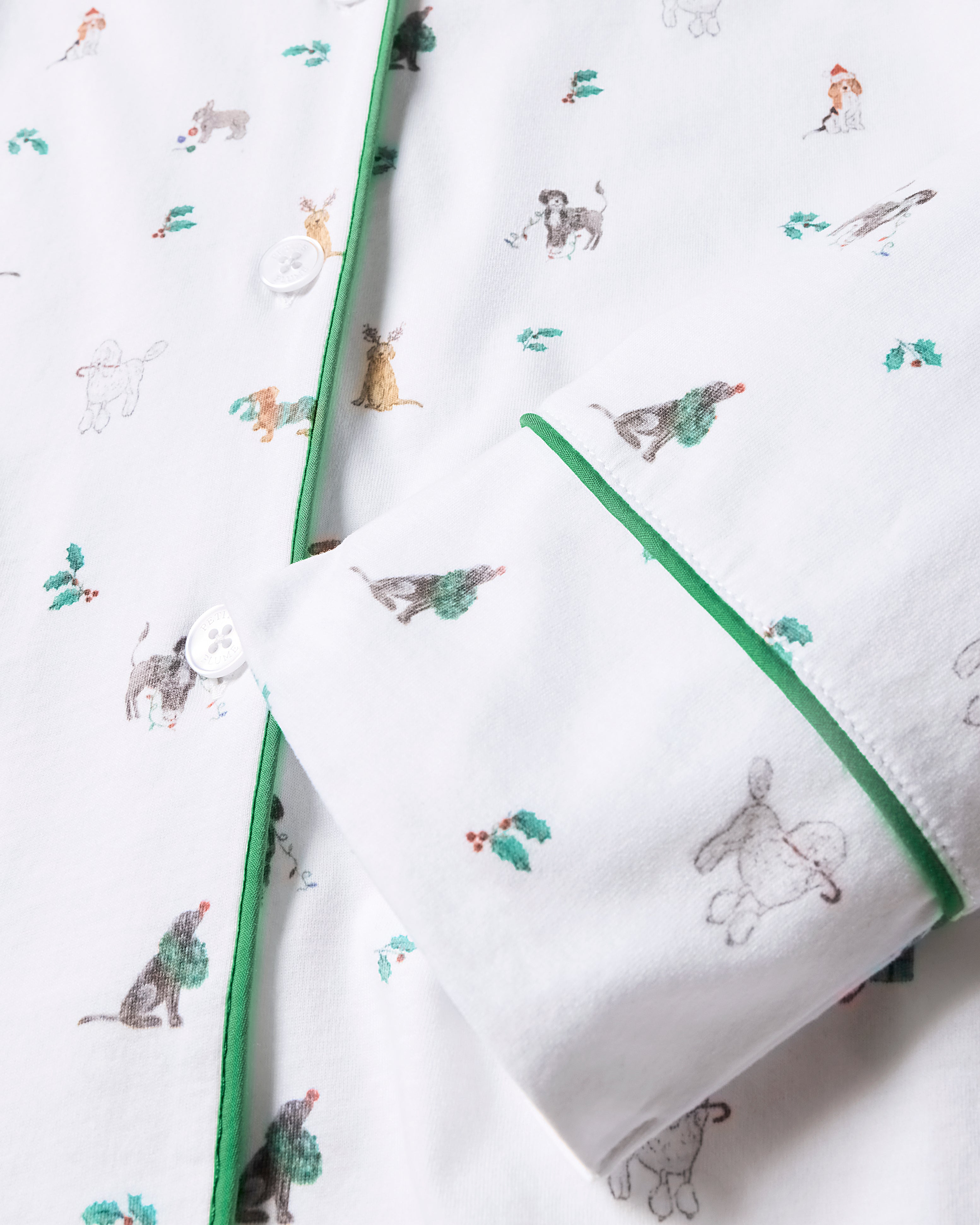 A close-up of Petite Plumes Womens Pima Pajama Set in Jingle Paws, an elegant white shirt with green trim featuring a festive dog and Christmas theme. Made from luxurious Pima cotton, it includes white buttons and a partially rolled sleeve for a relaxed holiday look.