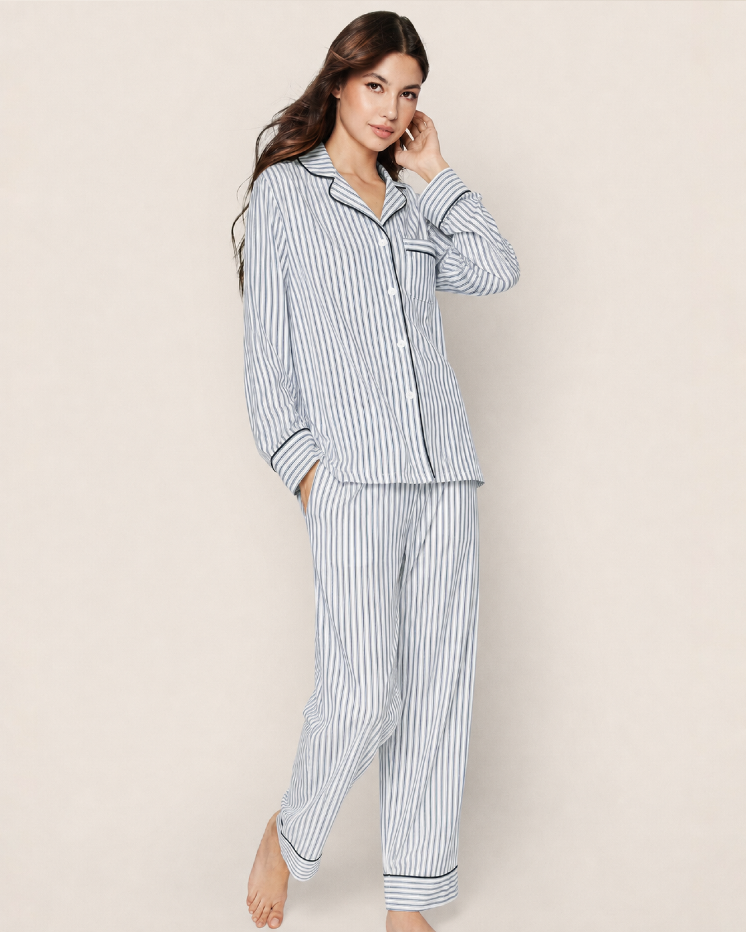 A woman stands barefoot against a plain beige background, wearing the Women’s Pima Pajama Set in Navy French Ticking, which features a matching long-sleeve top and pants with blue and white vertical stripes.