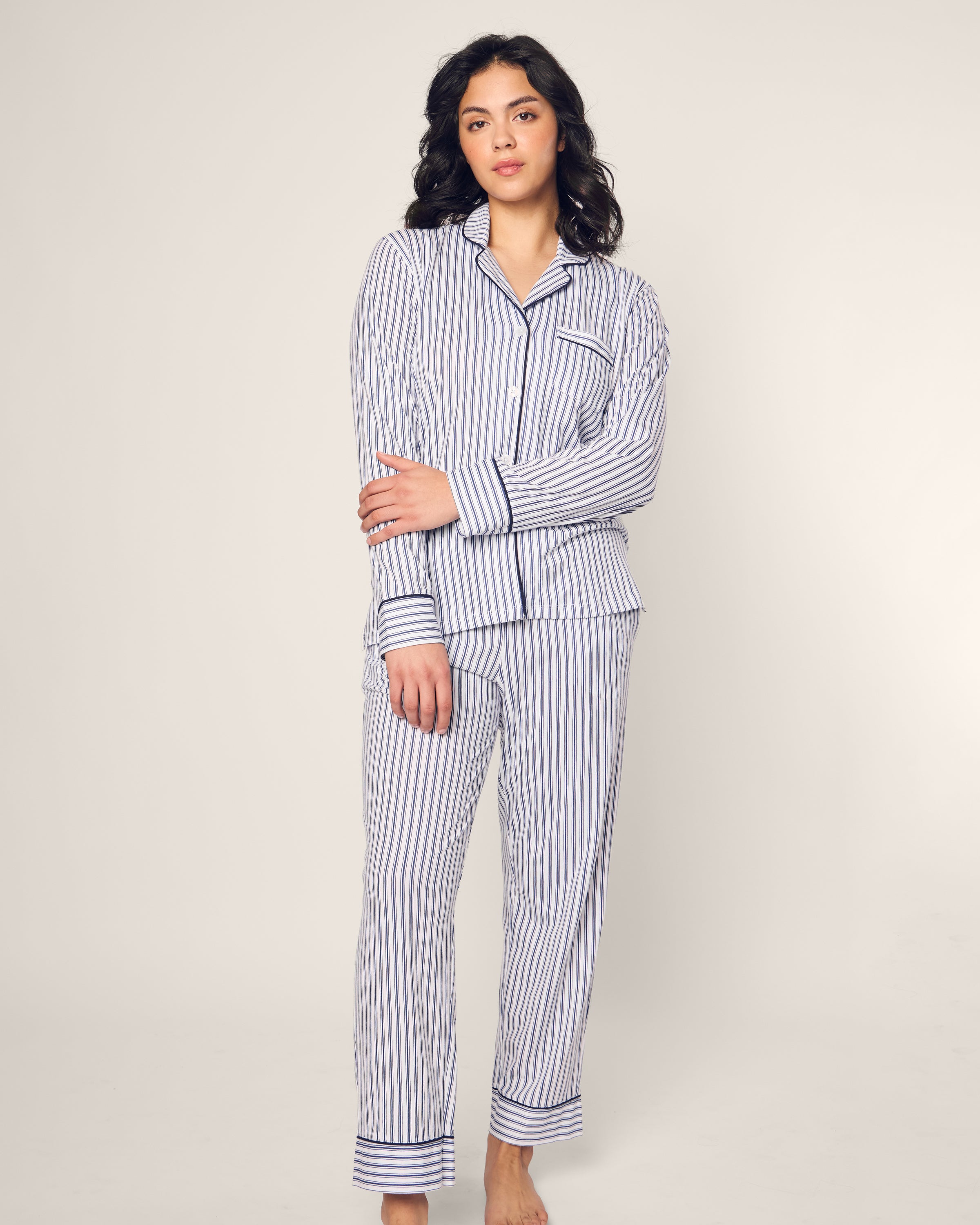 A person with long, dark hair stands relaxed against a neutral background wearing Petite Plumes Womens Pima Pajama Set in Navy French Ticking, crafted from soft Pima cotton, their arms crossed at the elbow.