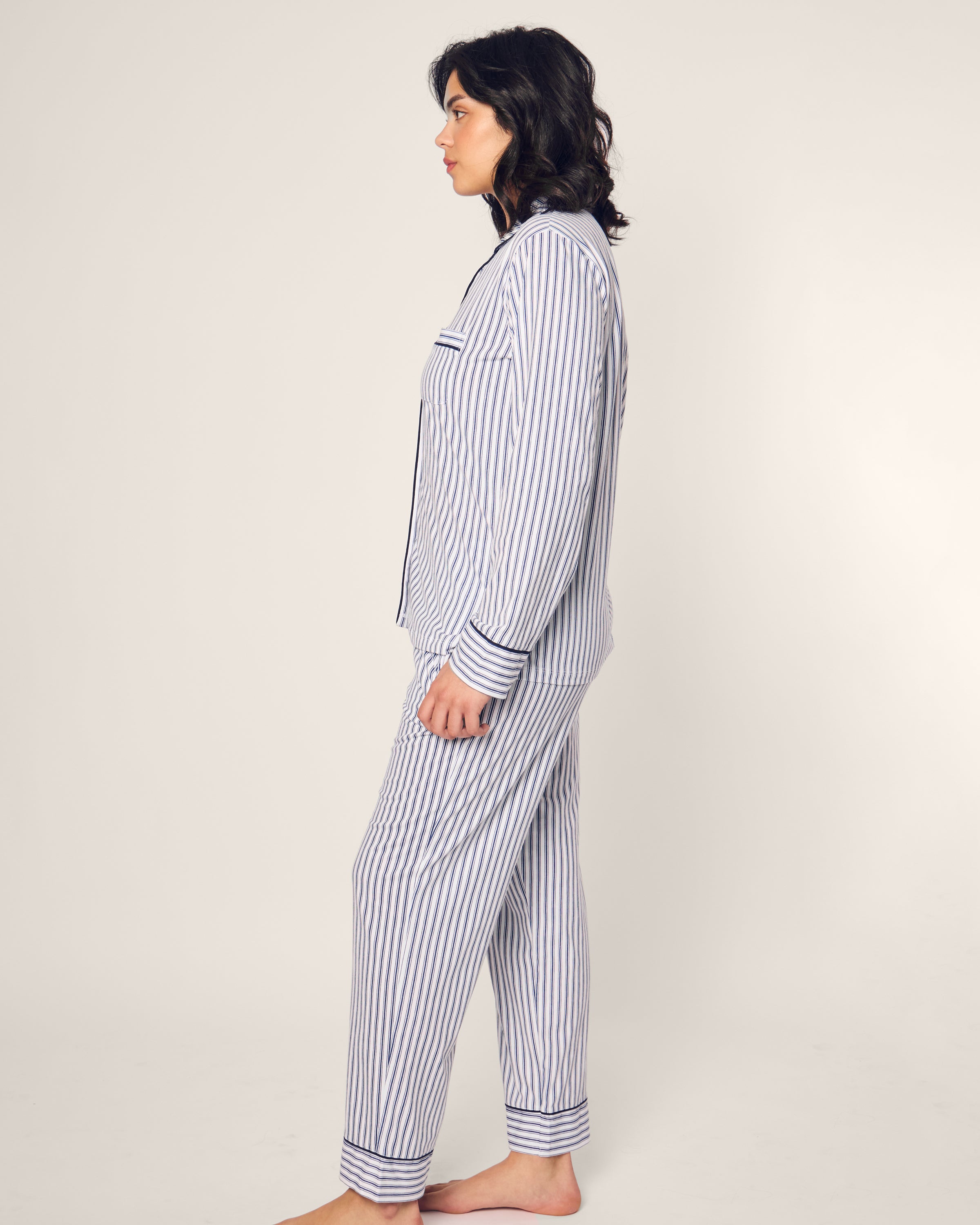 A person stands sideways in the luxurious Womens Pima Pajama Set in Navy French Ticking by Petite Plume, featuring dark horizontal and vertical stripes on a plain light background, crafted from exquisite Peruvian Pima cotton.