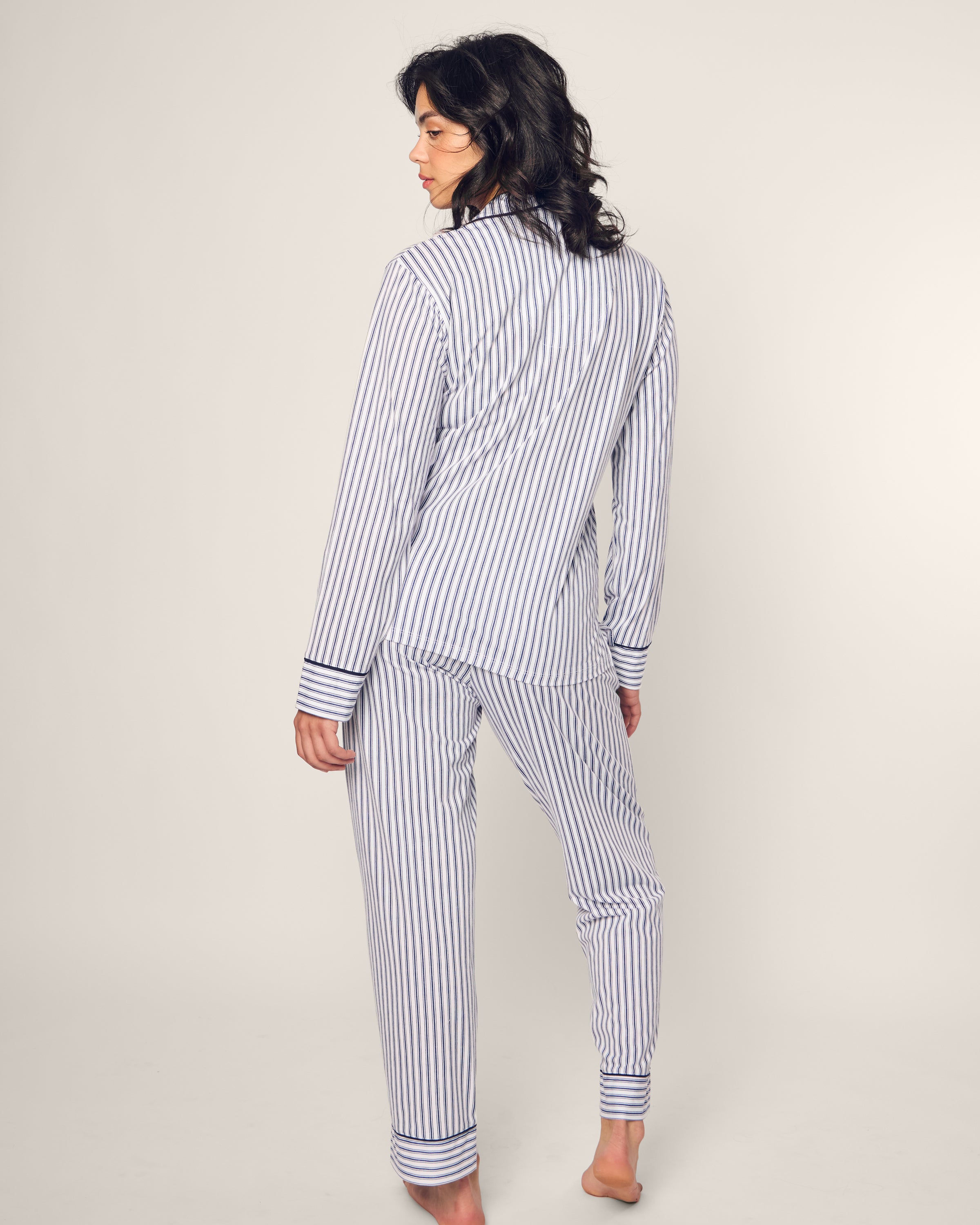 A person stands barefoot, facing away, wrapped in luxury sleepwear—a Womens Pima Pajama Set in Navy French Ticking by Petite Plume crafted from silky Peruvian Pima cotton. The design reflects classic French ticking elegance against a plain, light-colored background.